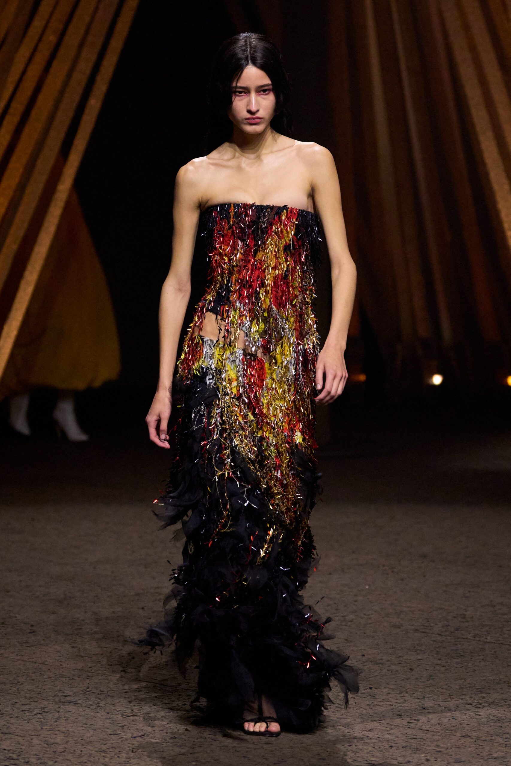 Strapless column gown veiled in red, gold and black tinsel fringe, hip slit showing through. Barely there black thong sandals. After the parade float is stripped, confetti still stuck to your ankles.