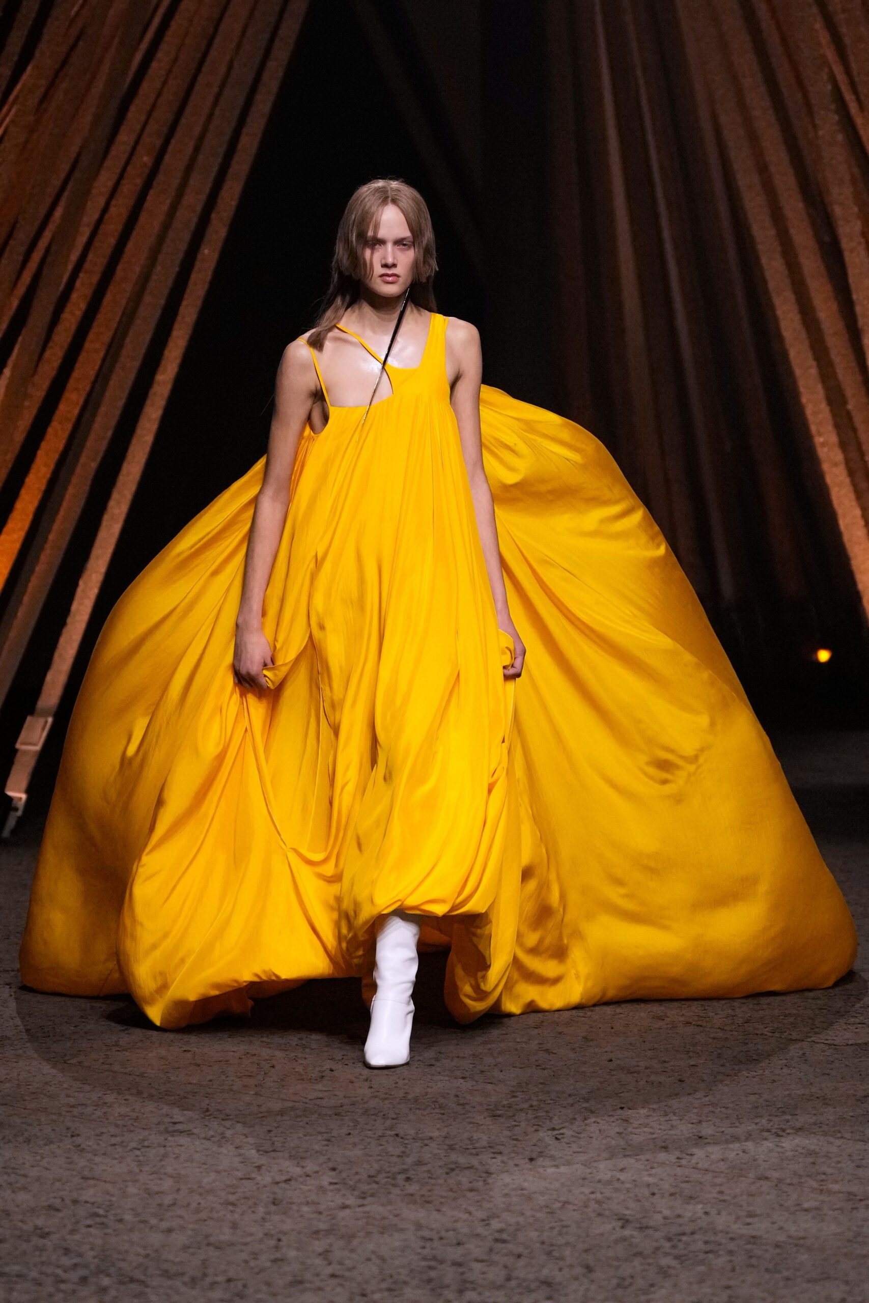 School-bus yellow gown cut with a skewed neckline and swollen train. White boots peeking from the pooled hem. Crossing a museum atrium with something too large to fit the lift.