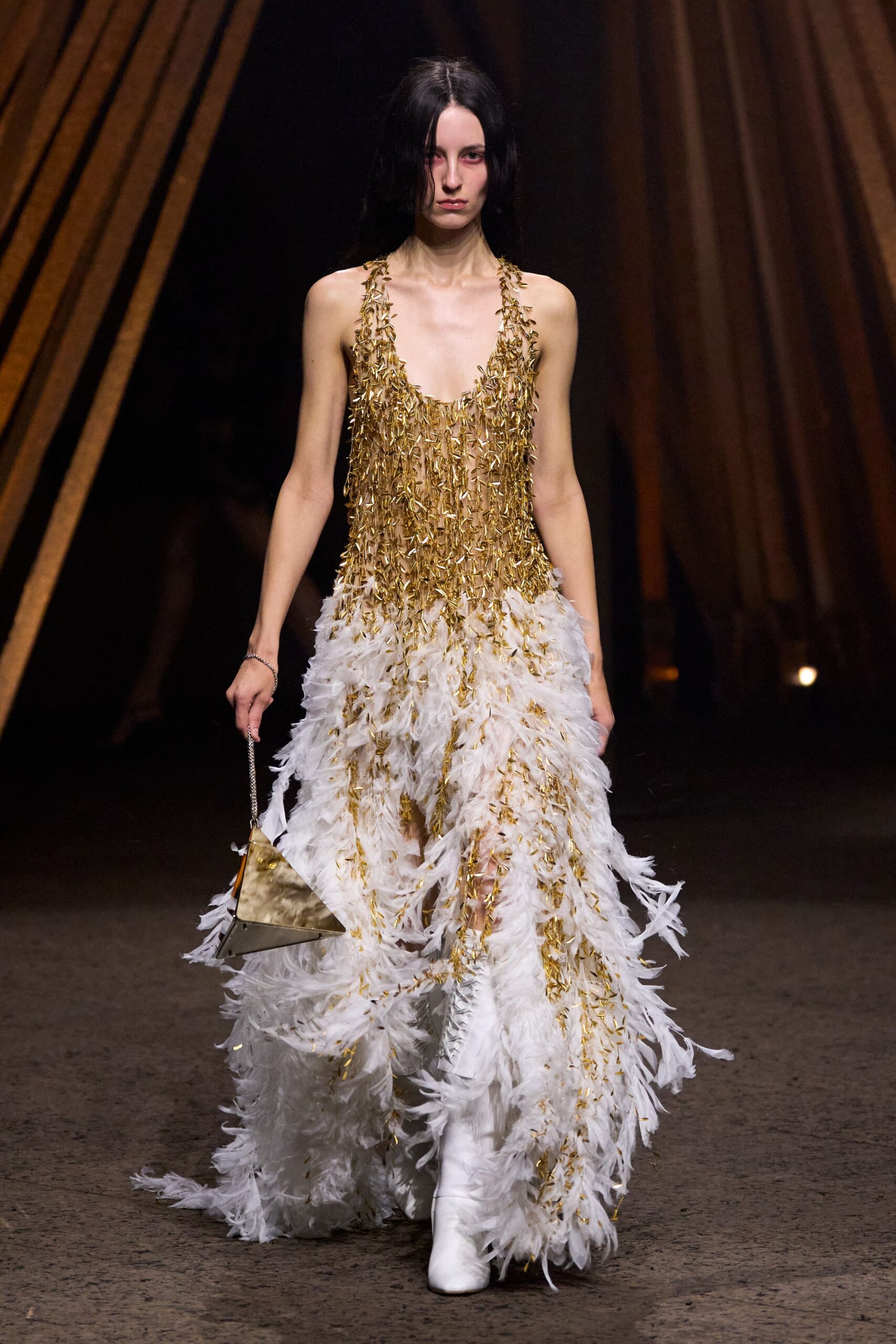 Plunging gold fringe gown dissolving into white ostrich-feather swirls at the floor. Small triangular gold bag on a fine chain strap. White ankle boots peeking through the feathered hem. Walking across a hotel ballroom carpet after midnight confetti has already been swept.