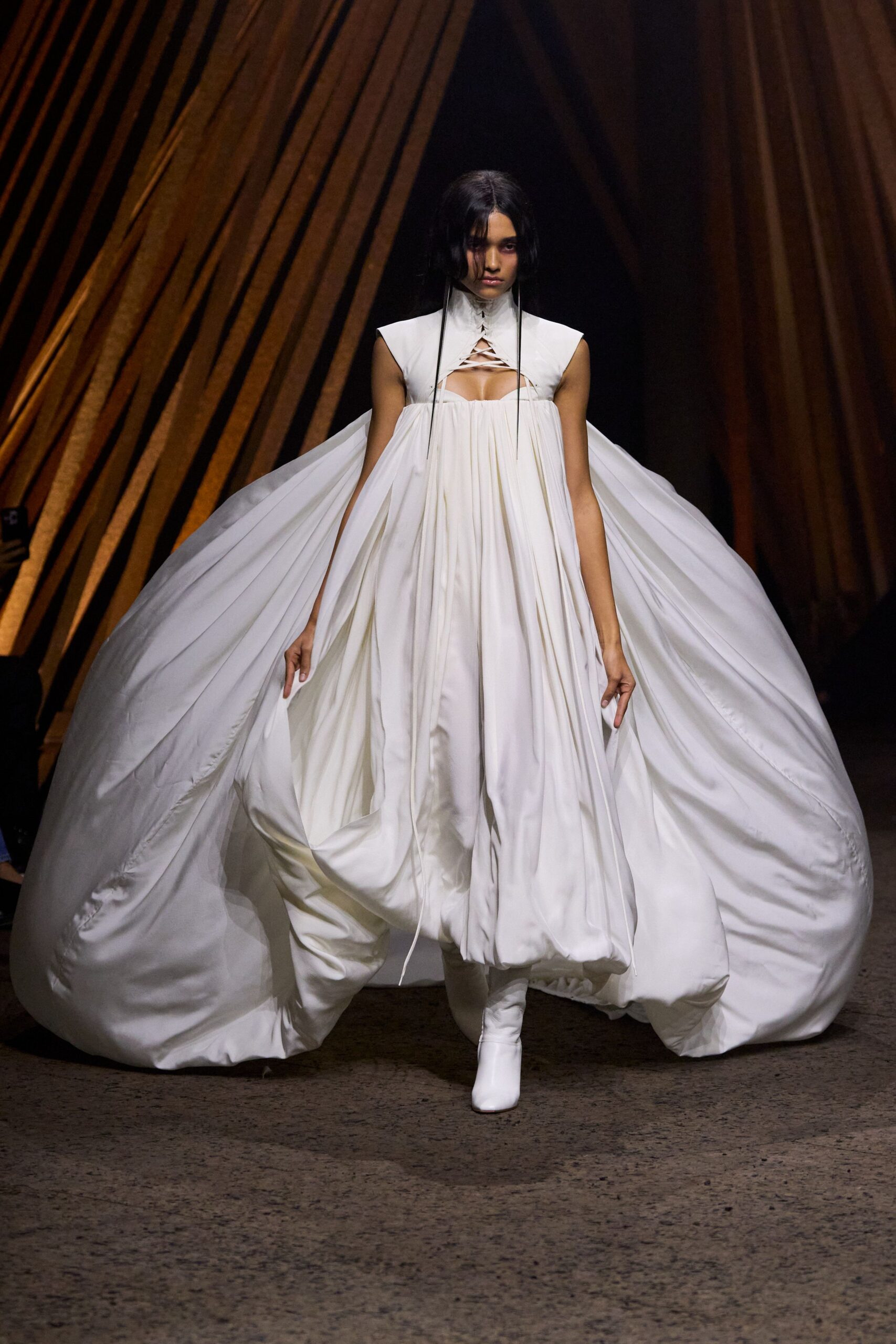 White high-neck dress with lattice lacing and a cutout over the bust. Immense gathered satin volume billowing from the sides, hem ballooned into pillows. White fitted boots, smooth and unbroken at the ankle. Crossing a marble hall while every door stands open to the wind.