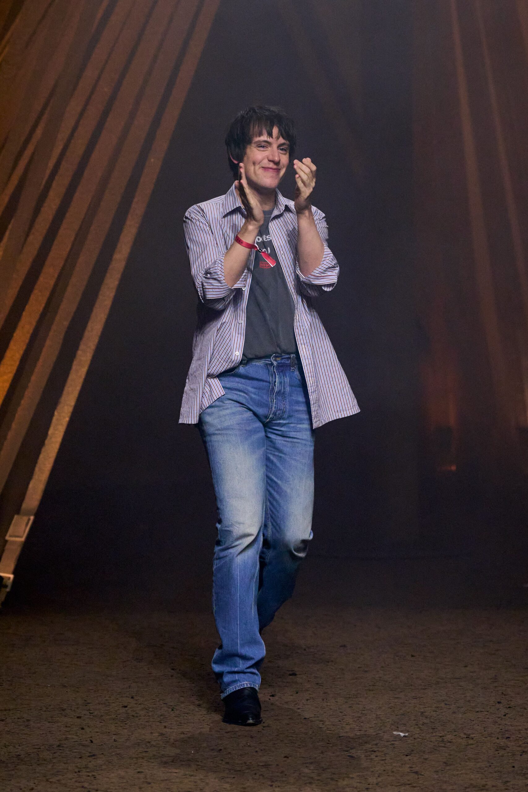 Blue and white banker-stripe shirt worn open, sleeves pushed to the forearm. Faded charcoal graphic tee peeking through the placket. Light-wash bootcut jeans with whiskering and a soft break at the shoe. Black leather square-toe shoes. Taking applause in a school auditorium after the amps finally stop humming.