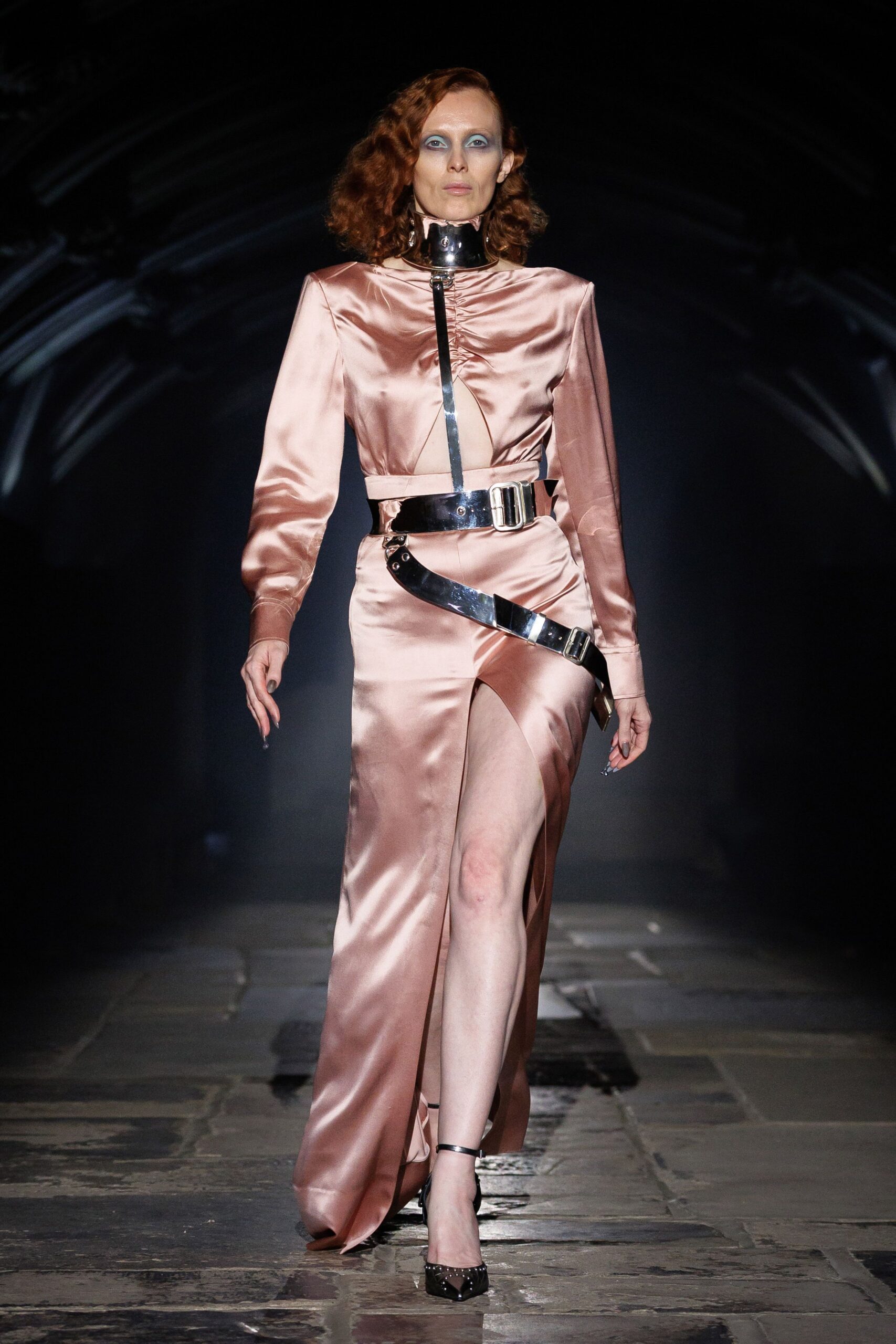 Mirror-metal choker collar fastening high under the jaw. Blush satin column gown, ruching pinched tight beneath a metal strip. Black patent straps buckle at the waist and slice diagonally over the hip. Black patent pumps with a pointed toe and ankle strap. Crossing a museum nave after midnight, floors still cold from the day.