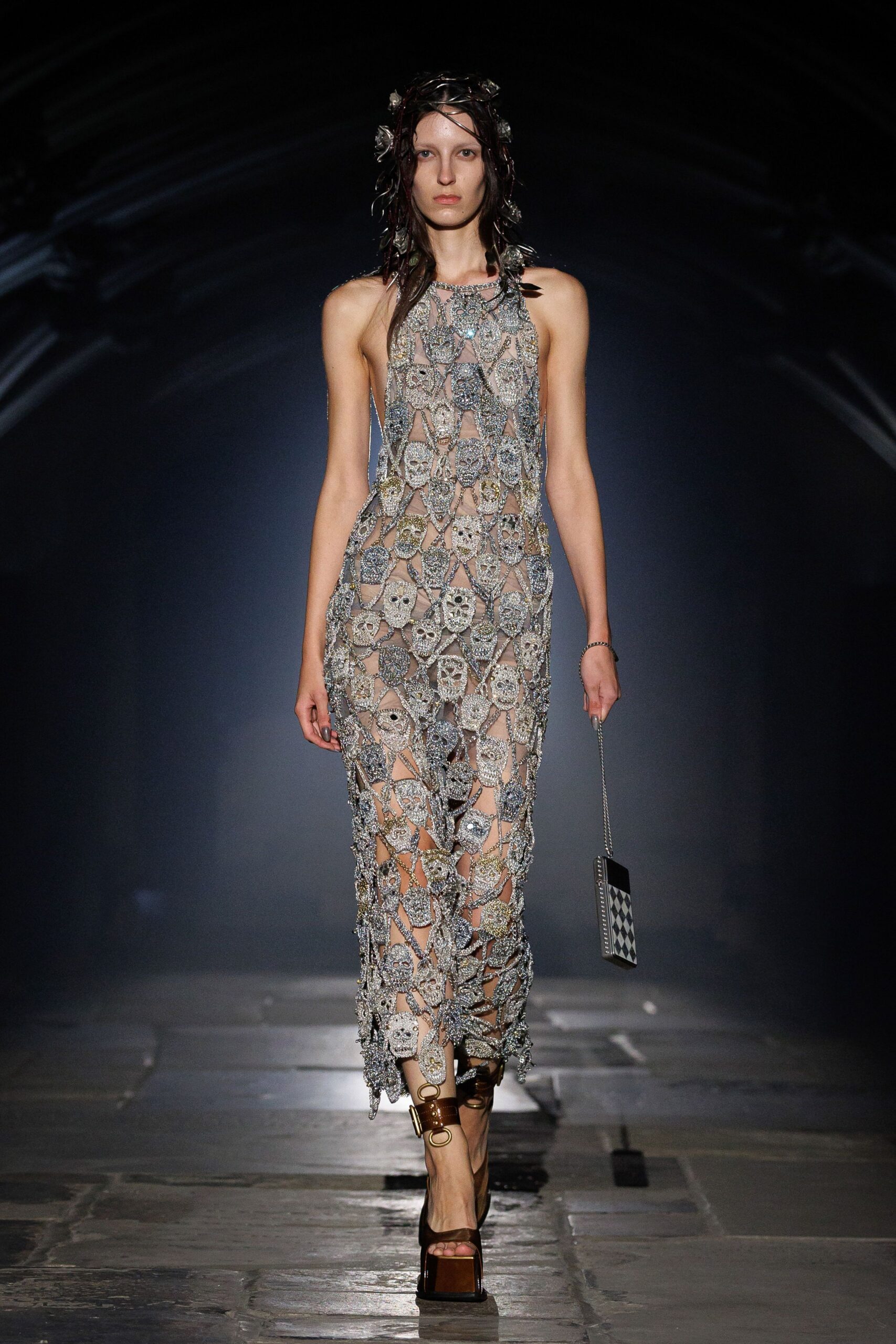 Nude column dress netted with silver beaded skulls, open between every motif. Dark cords tangled with crinkled foil flowers. Slim phone case on a chain, harlequin faceplate. Brown patent platforms with oversized ring hardware at the ankle. After midnight at a funfair, prizes gone and the lights still humming.