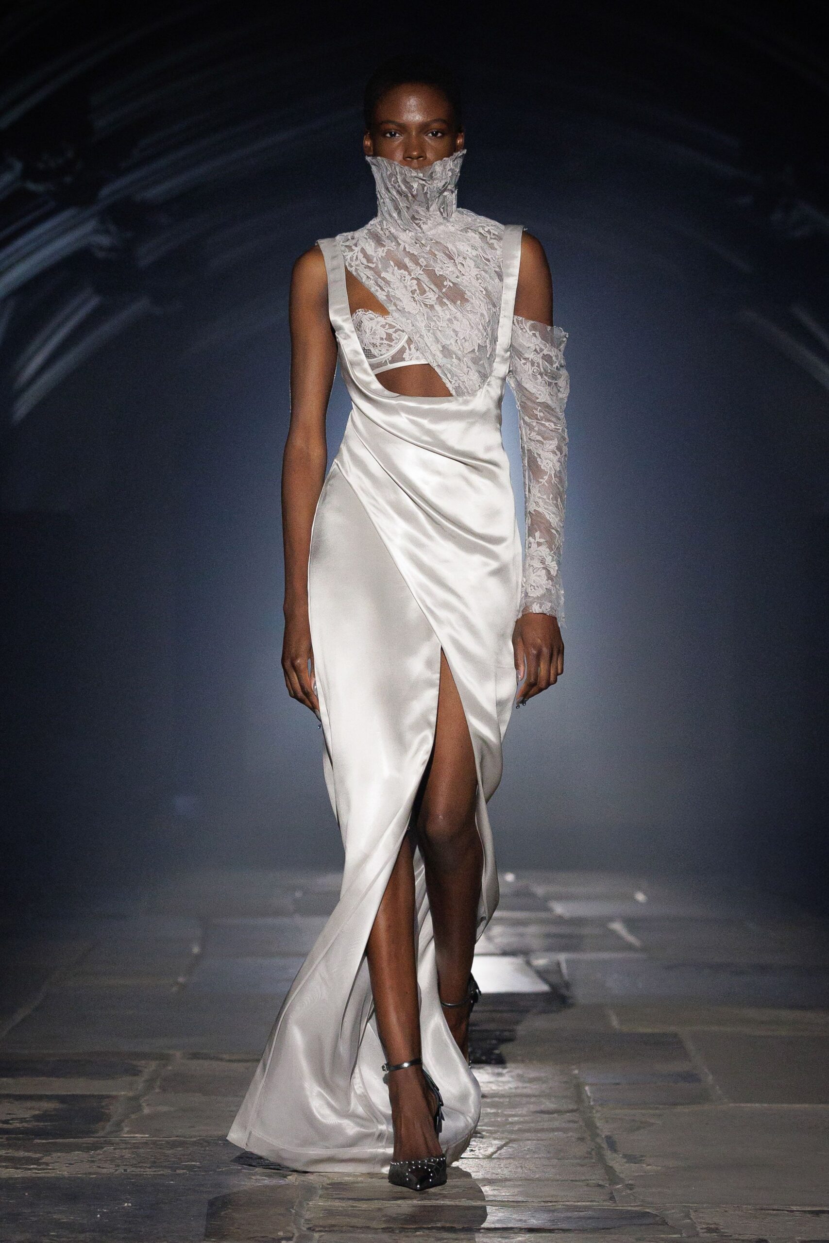 Ivory satin gown draped low at the bust, split high in front. Sheer floral lace wrapping the neck and one arm, bra exposed. Black pointed heels with tiny crystal studs at the toe. Registry office stairs, bouquet paper going soft in your hand.