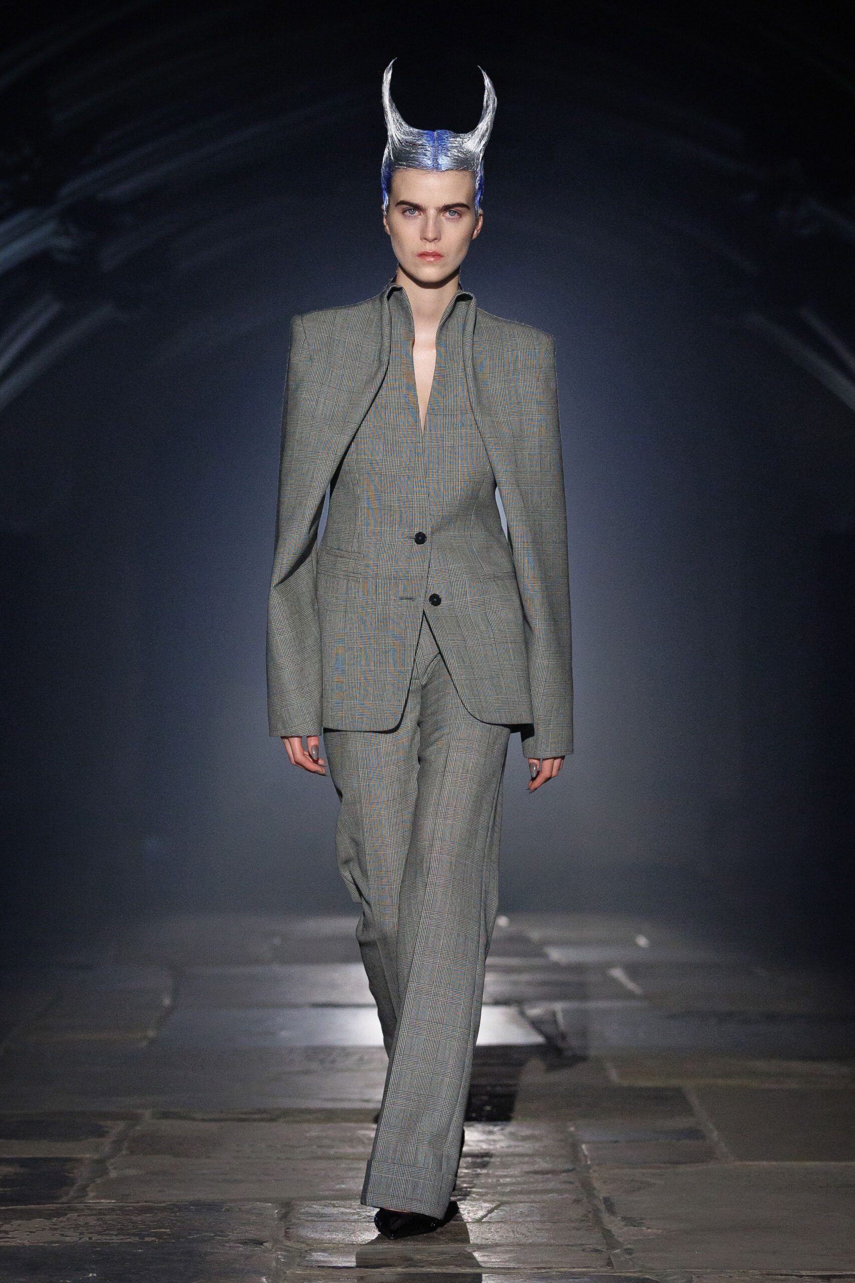 Silver sculpted horns rising from a slicked cap of hair. Grey Prince of Wales blazer with knife-sharp shoulders and two black buttons. Matching wide-leg trousers breaking heavily over the shoe. Crossing a cathedral floor before anyone else has unlocked the doors.