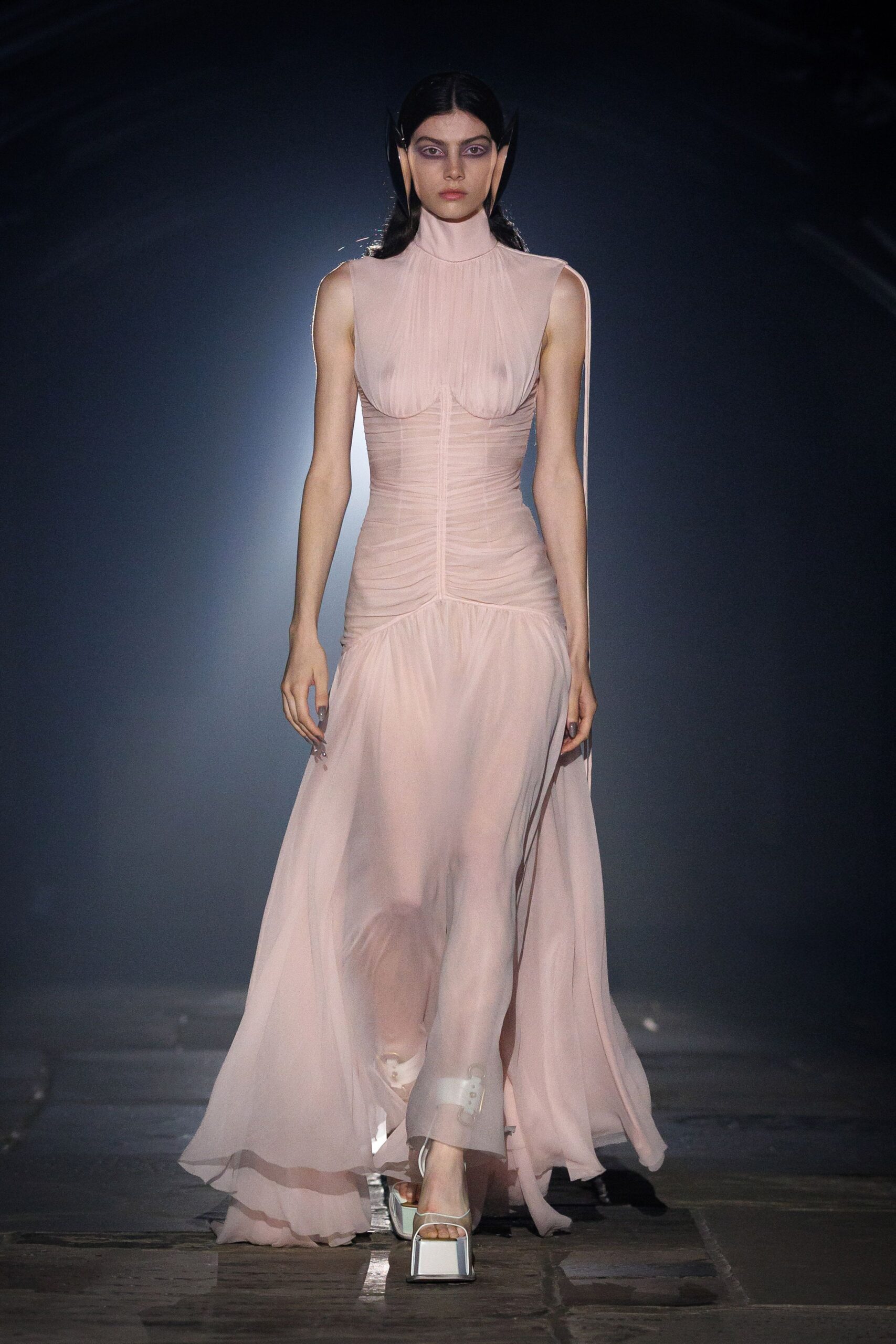 Blush chiffon gown, ruched through the torso with sheer gathered cups. Sharp black fins bracketing the head. White platform sandals with a thick square sole. Crossing a museum atrium just after the fountains switch off.