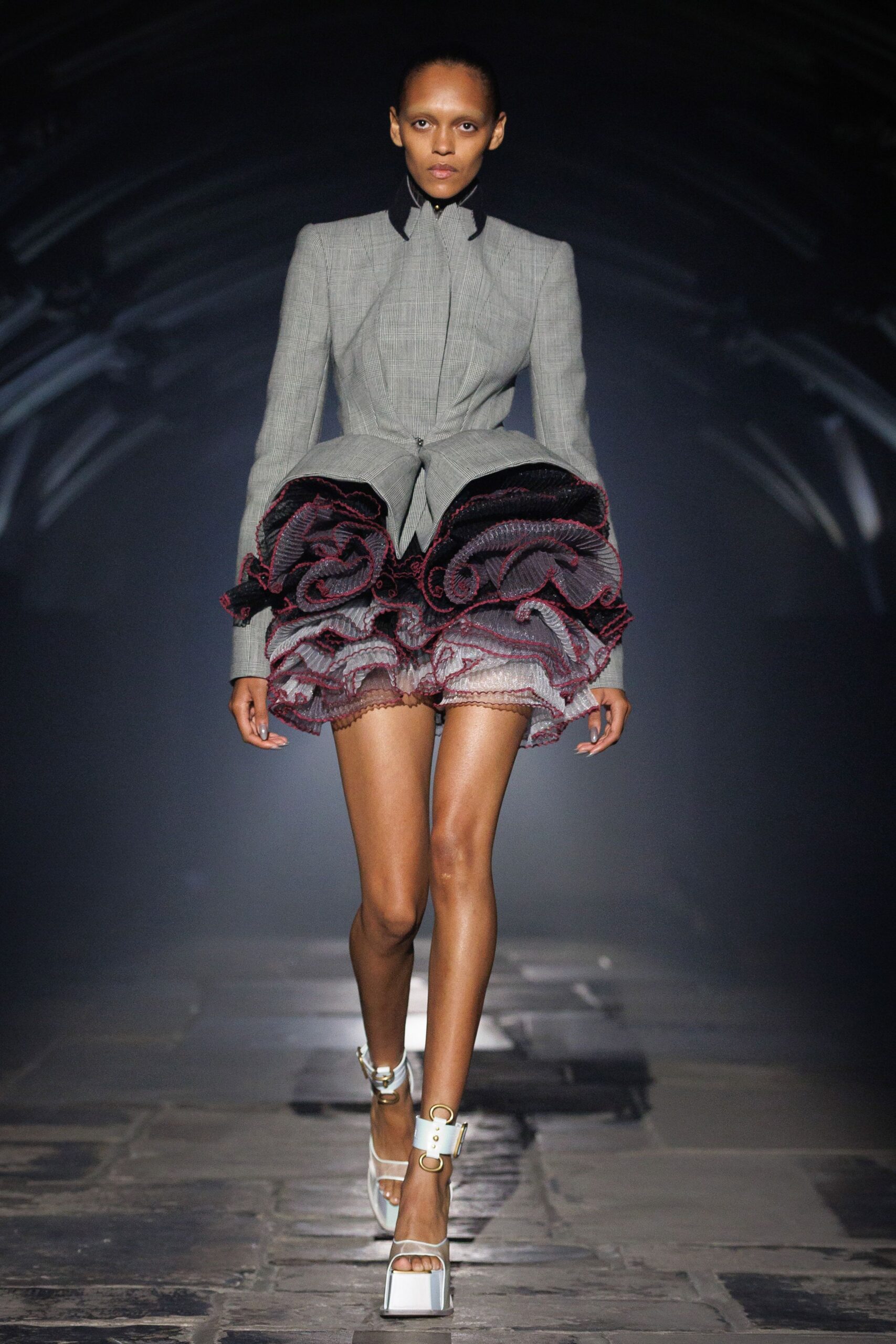 Grey prince-of-wales jacket, waist pulled tight above a split peplum. Plum-edged organza ruffles boil out in dense spirals from the hip. White platform sandals with broad straps and oversized gold O-rings. Crossing a museum atrium while the string quartet is still tuning.