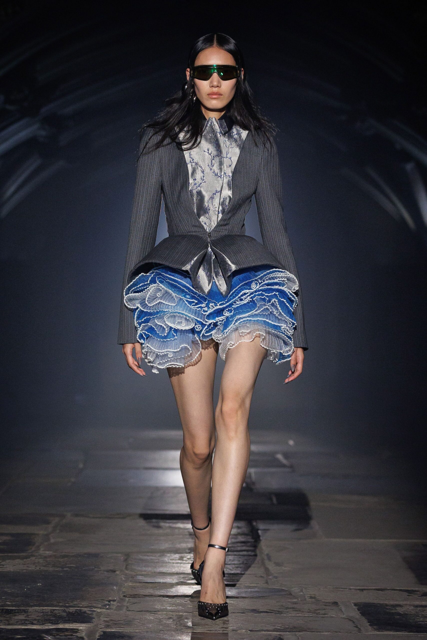 Single smoked shield lens with a green flash across the brow. Charcoal ribbed jacket, zip-fastened and kicked into a sharp peplum. Silver satin blouse printed with cobalt trailing branches. Tiered blue mesh ruffles edged in white picot stitching. Black patent ankle-strap pumps. Crossing the museum atrium after the rain stopped.