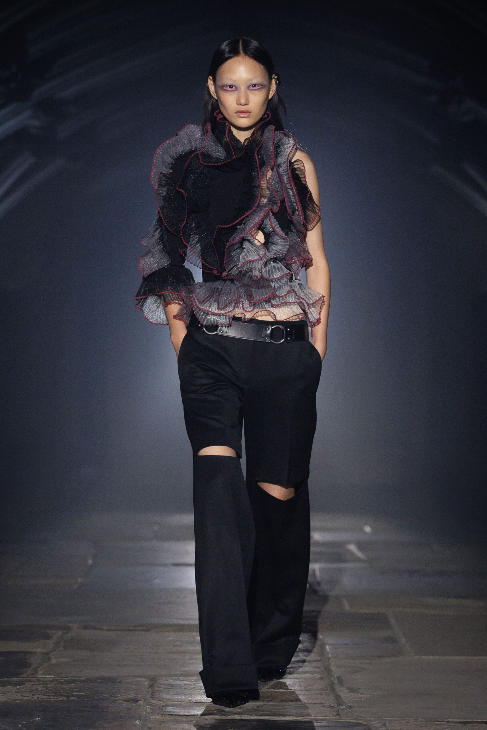 Asymmetric black mesh ruffles edged in wine over silver accordion pleats. Black tailored trousers sliced open at both knees. Wide leather belt with an oversized D-ring buckle. Pointed black shoes, barely flashing under the hem. Crossing a stone courtyard after midnight rain, still carrying the argument.