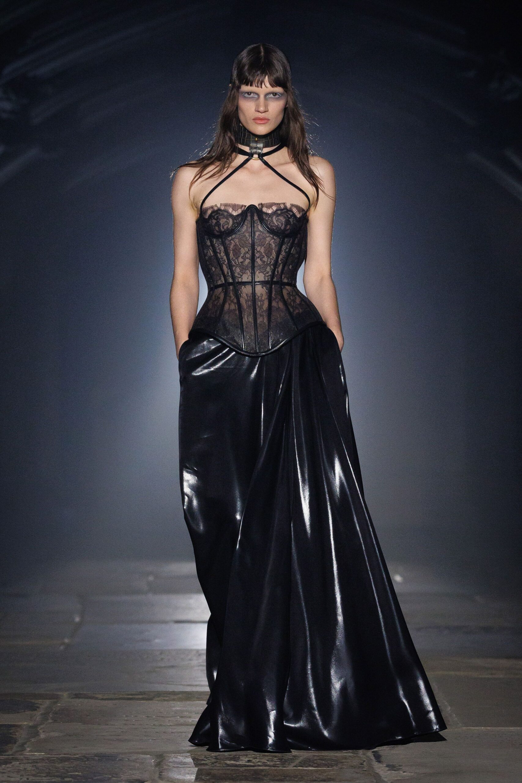 Black leather choker harness with a brass ring at the throat. Sheer black lace corset, boned seams curving past a dipped basque hem. Floor-length black skirt in high-shine coated fabric, pooling into heavy folds. Whatever you wear home when the hotel corridor still smells of smoke.
