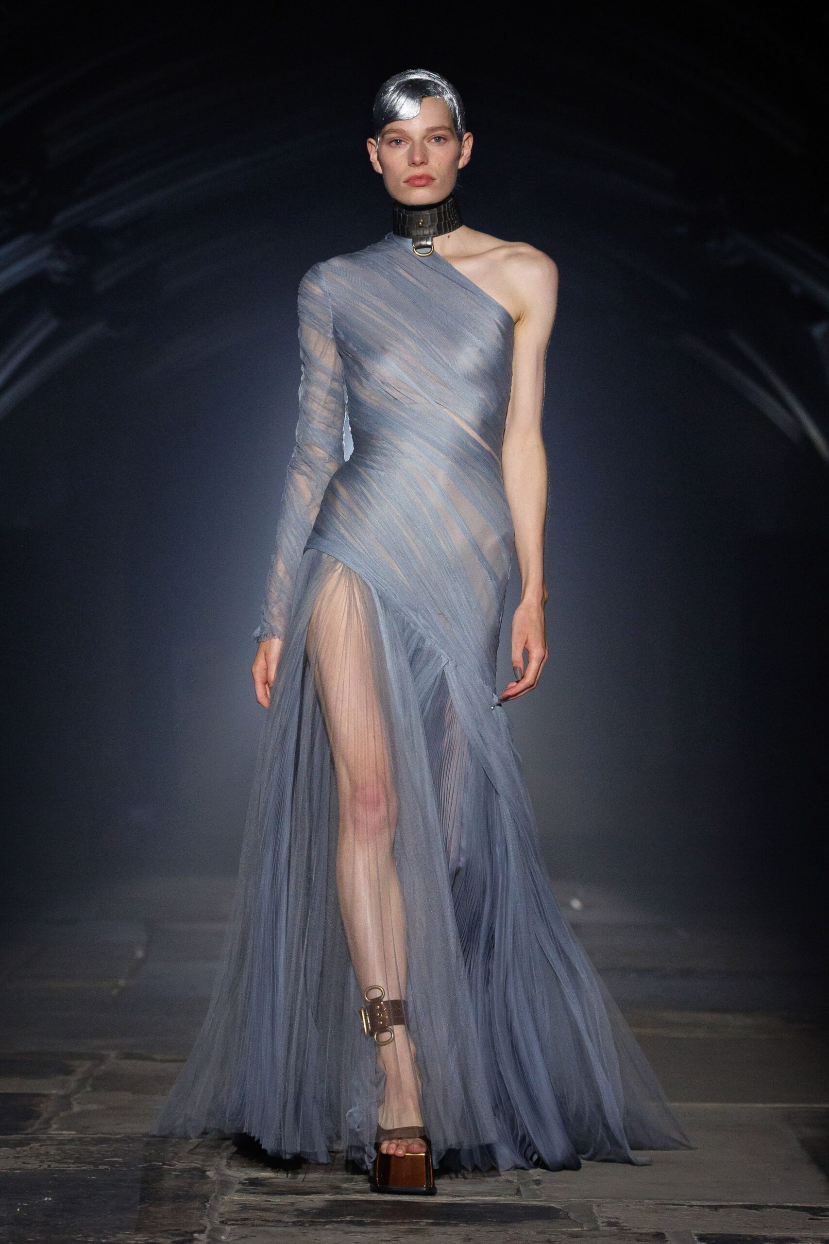 Black leather choker ringed with a brass O at the throat. Slate tulle gown spiraled one-shoulder, one sleeve sheer and the thigh split draped open. Platform sandals with brown leather ankle straps and brass rings. Crossing a museum courtyard after rain, hem drinking from the stones.