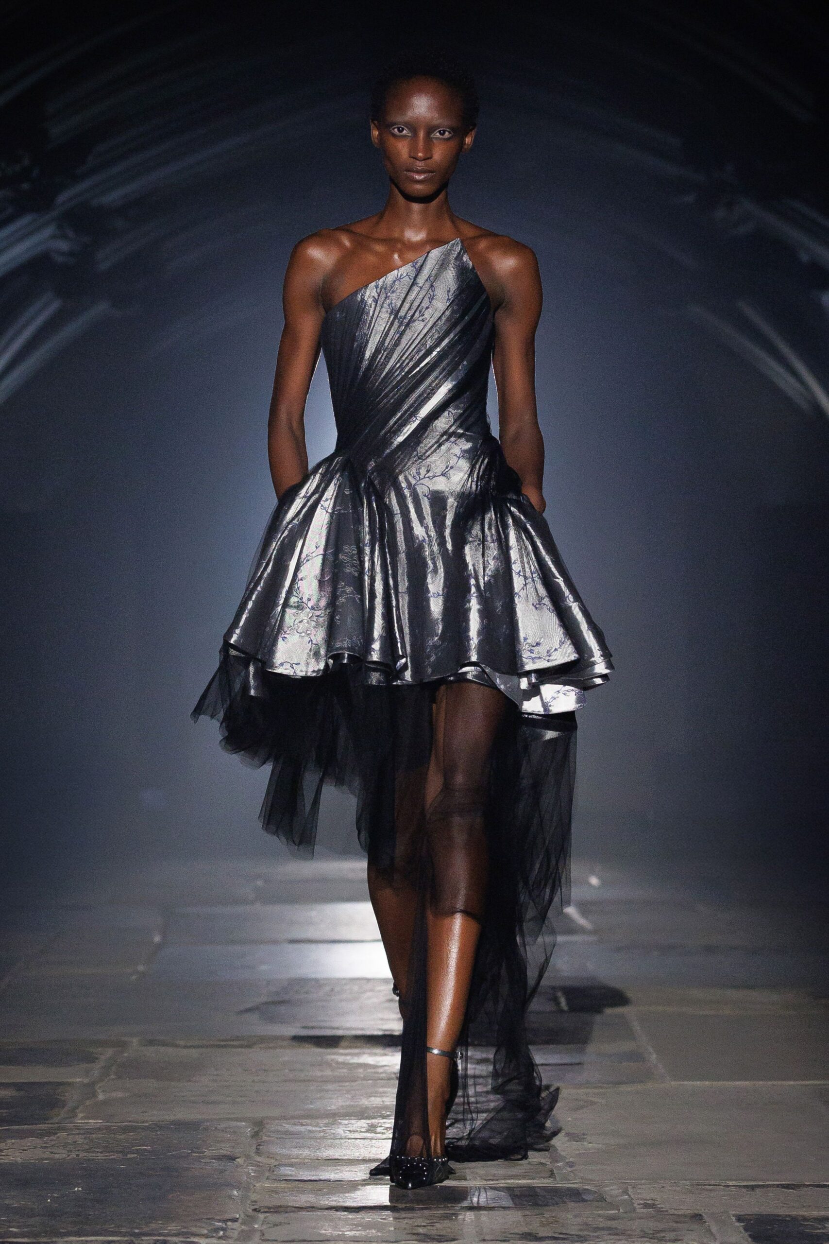 One-shoulder silver brocade dress, diagonally ruched and blown into panniered hips. Black tulle underskirt falling away in a sheer uneven tail. Black pointed heels with tiny studs across the toe. Crossing a stone courtyard after midnight, carrying your skirt out of the fountain spray.