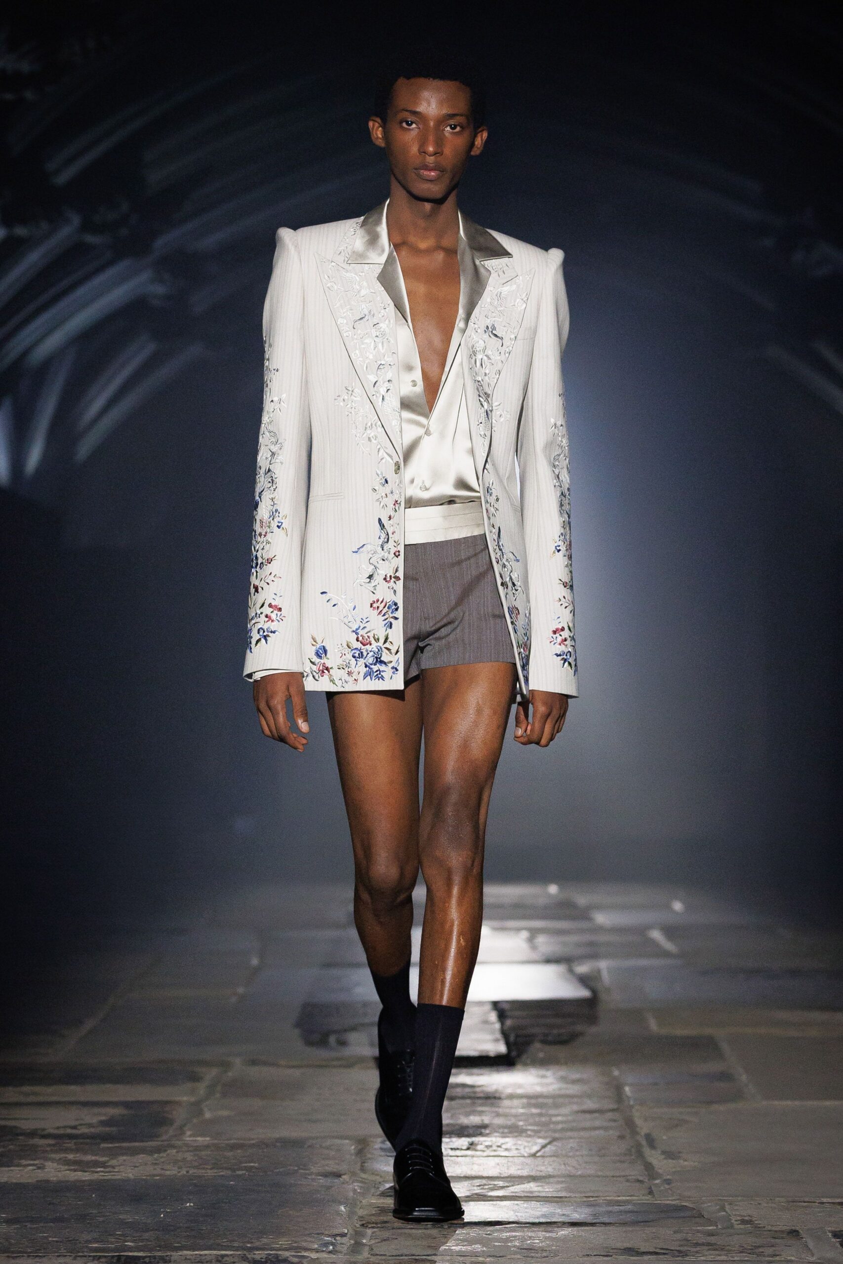 Ivory pinstriped blazer with padded shoulders and scattered floral embroidery. Pale satin shirt worn plunging, the third button catching light. Taupe tailored shorts under a wide cummerbund waistband. Black ankle socks. Square-toe black lace-ups. Crossing a stone courtyard after midnight mass, flowers crushed in one fist.
