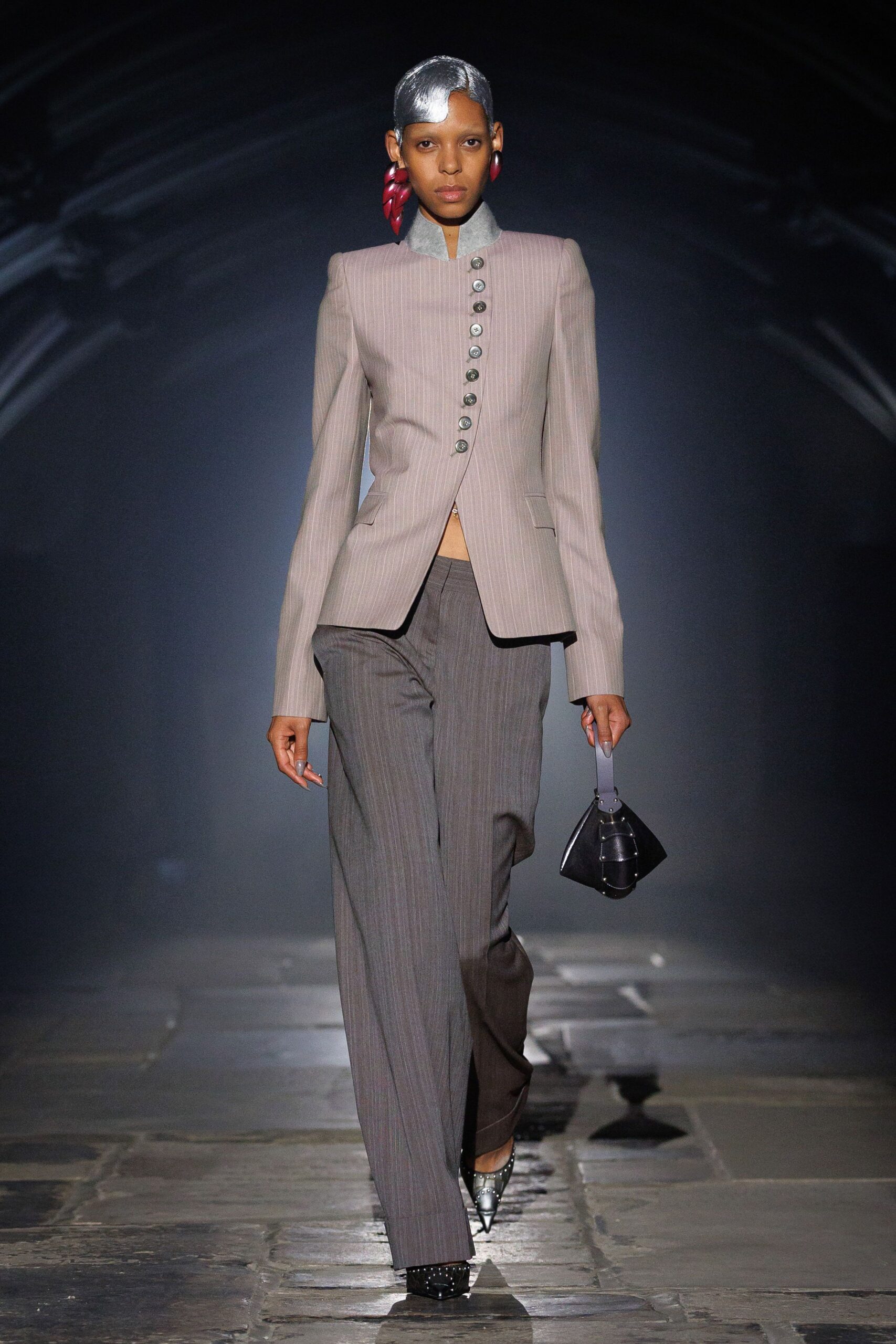 Blush pinstripe jacket with a pale felt stand collar and off-center pewter buttons. Charcoal pinstripe trousers cut wide through the leg. Glossy cherry-red drop earrings. Small black leather bucket bag with layered flap panels. Studded silver cap-toe pumps. For crossing a museum atrium while rain dries on the stone.