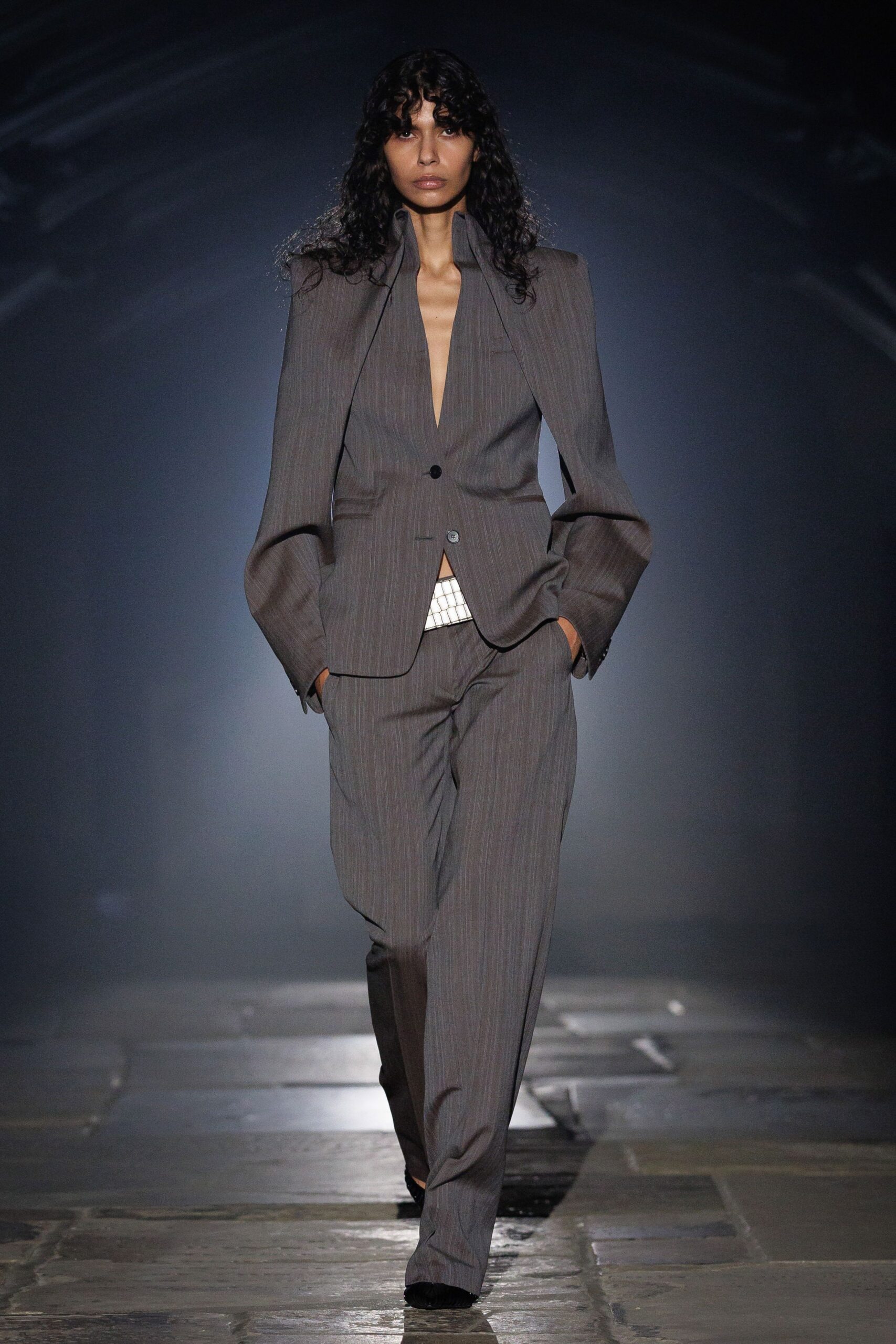 Taupe pinstripe blazer with razor shoulders and a plunging split stand collar. White grid-textured waistband peeking through the unfastened hem. Matching straight trousers, softly pooling over the shoes. For the conference call you take from a half-empty hotel lobby.