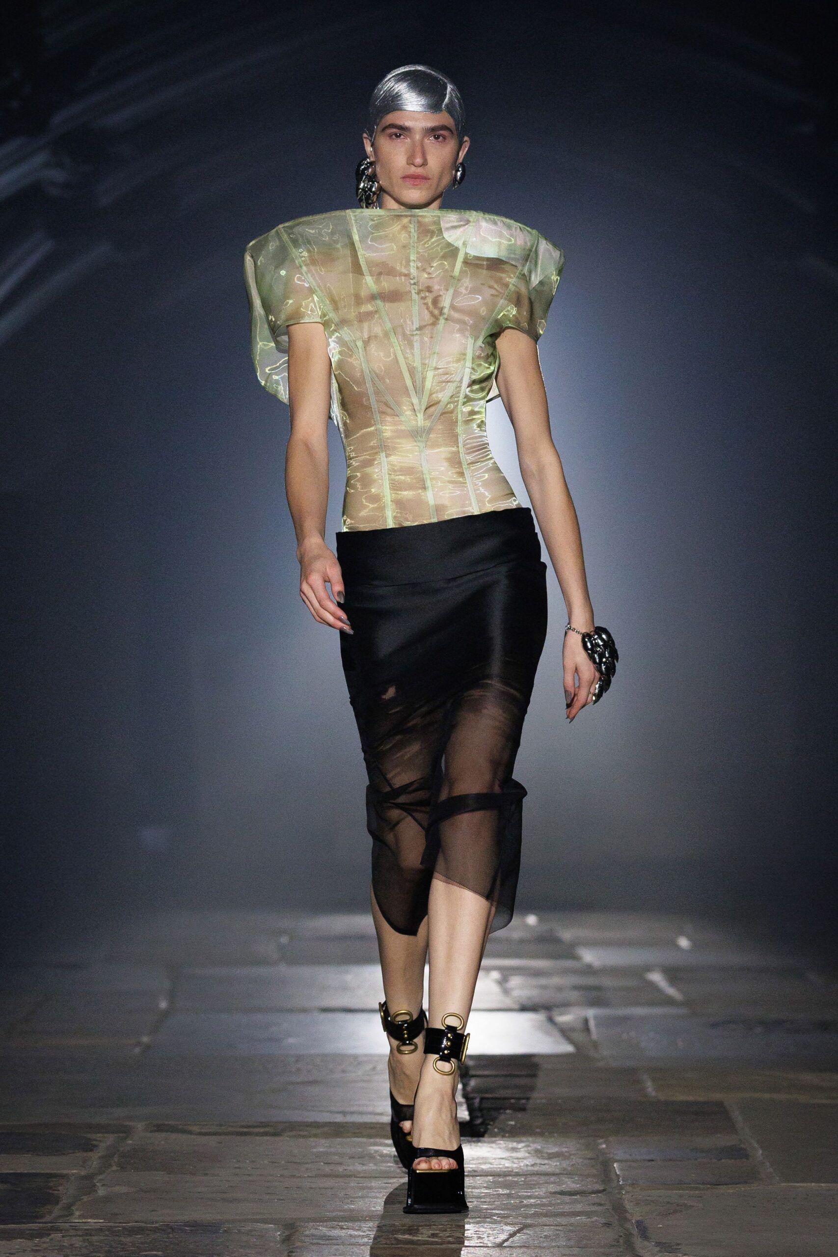 Iridescent organza top, corset seams pinning a sheer nude underlayer. Black chiffon tube skirt, one panel slipping crooked across the knees. Chunky black chain-link hoops. Matching black chain cuff. Patent platforms with brass rings buckled at the ankle. Crossing a museum atrium after rain, floor still shining underfoot.