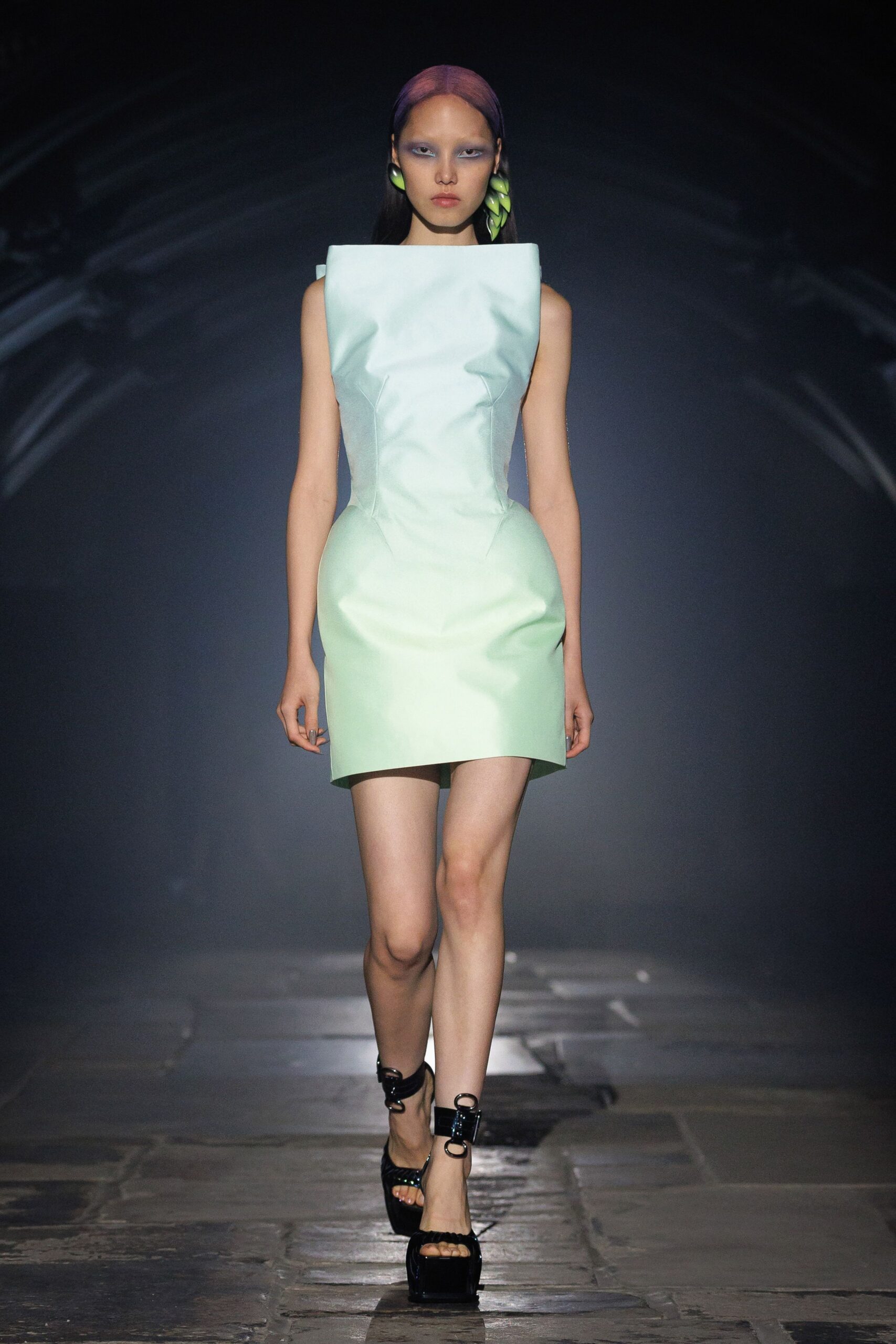 Mint shift dress in stiff satin, squared neckline jutting from the shoulders. Pale green petal-drop earrings stacked like folded leaves. Black patent platforms with oversized ring buckles at the ankles. Crossing a conservatory after dark, concrete still warm underfoot.