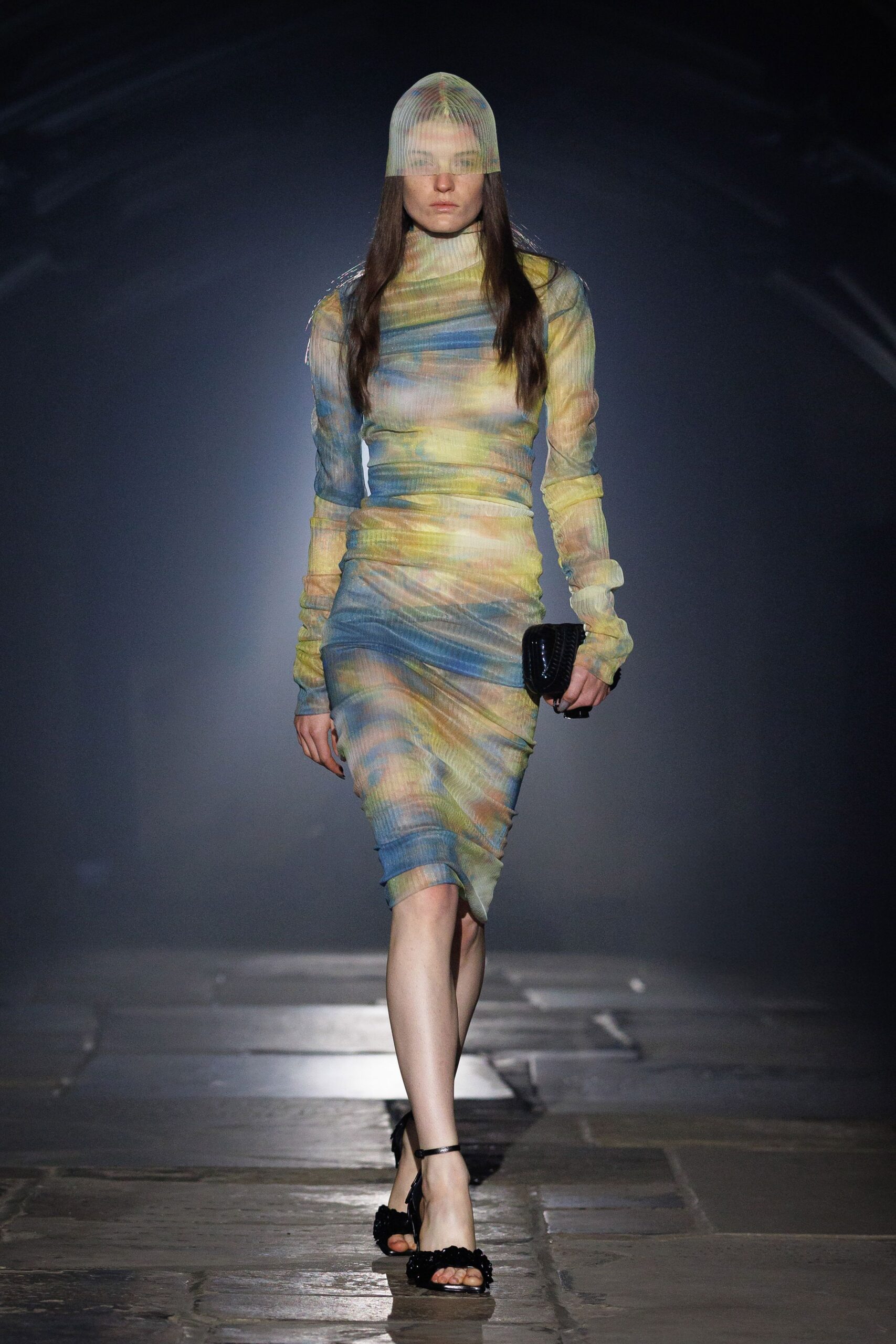 Fine ribbed mesh cap dropping straight over the eyes. Sheer ruched midi dress in watery pastel bands, sleeves pooling at the wrists. Glossy black woven clutch tucked hard under one arm. Black ankle-strap sandals with shaggy rosettes across the toes. Crossing a museum courtyard just after the rain machine shuts off.