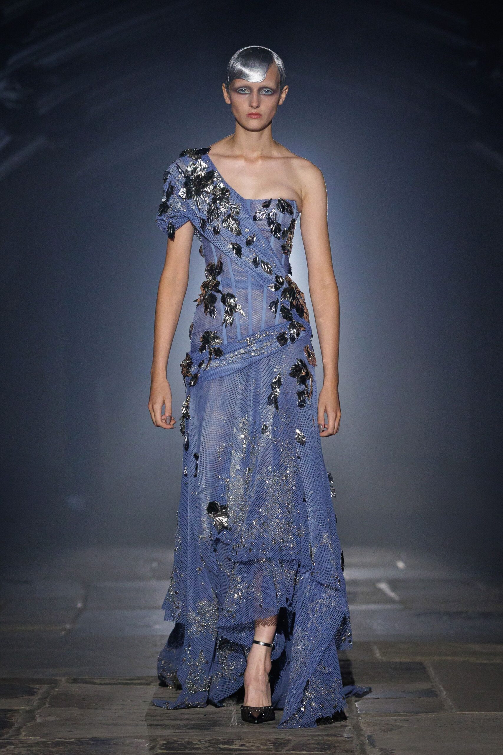 Silver molded skullcap dipping into a widow's-peak point. Periwinkle open-mesh gown, one-shouldered and corseted, strewn with crinkled foil petals. Black patent pumps with a slim ankle strap. Crossing a museum atrium after the gala, confetti still stuck to the floor.