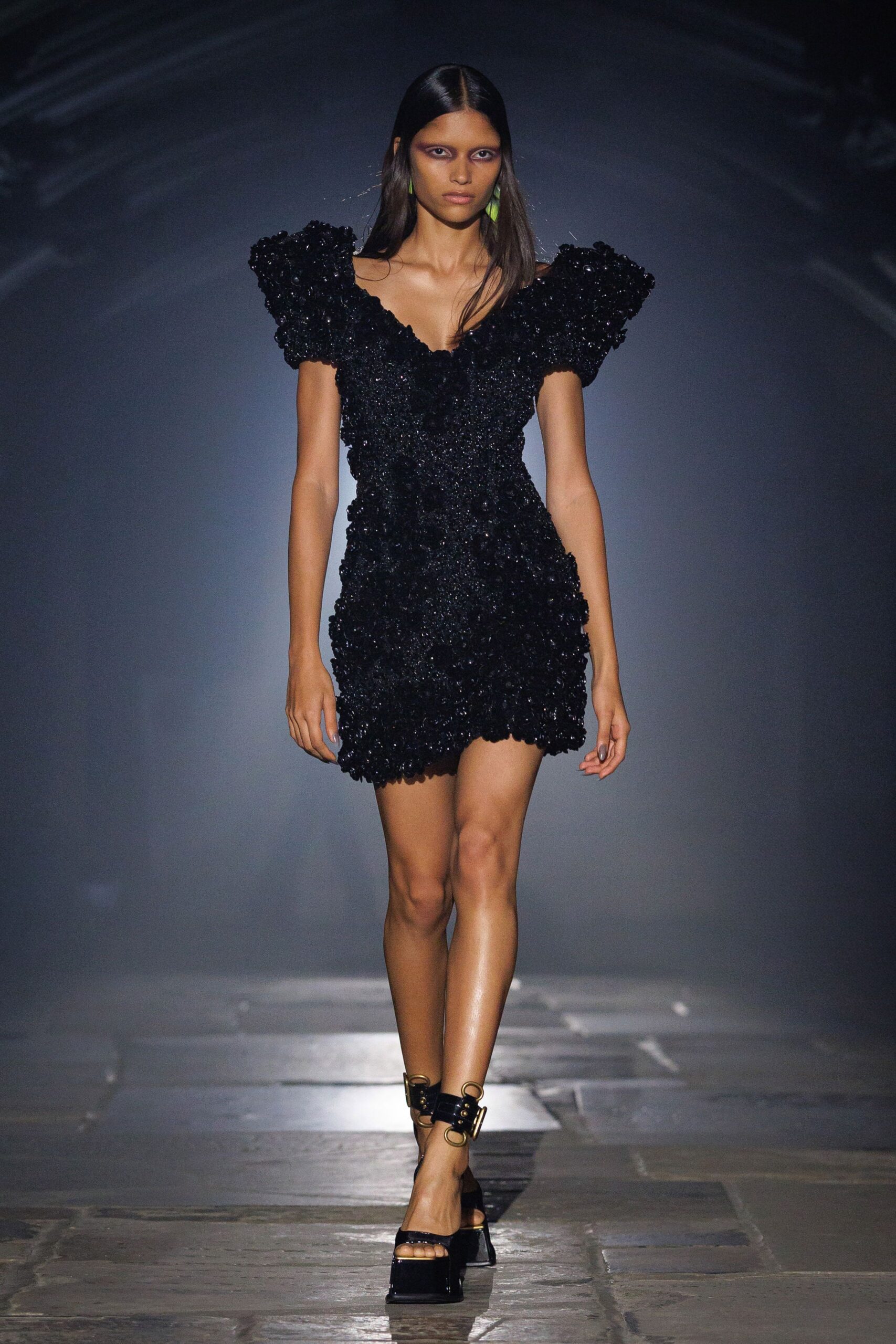 Black textured mini dress crusted with paillette-like knots, shoulders jutting sharply. Small acid-green drops at the ears. Black patent platforms with oversized gold ankle rings and a block heel. Crossing a museum courtyard after rain, when the stone still throws back light.