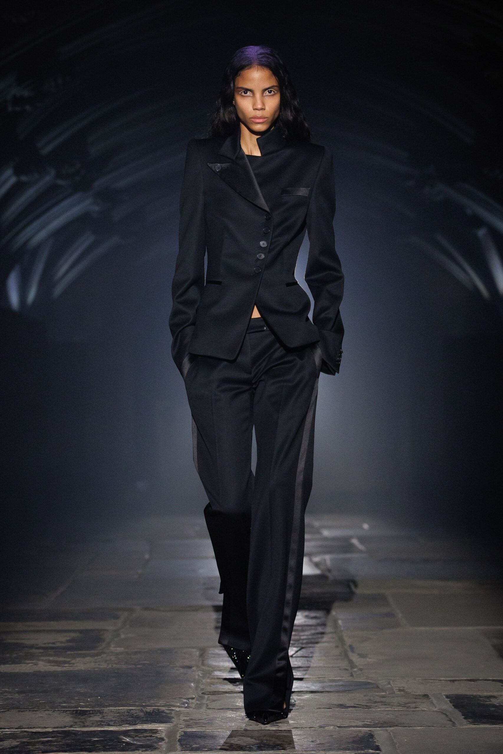 Black tailored jacket with satin shawl lapel and a crooked row of buttons. Black straight trousers with satin side bands and a low sliver of midriff. Pointed black boots peeking from the hem. Crossing a stone cloister after midnight mass let out.