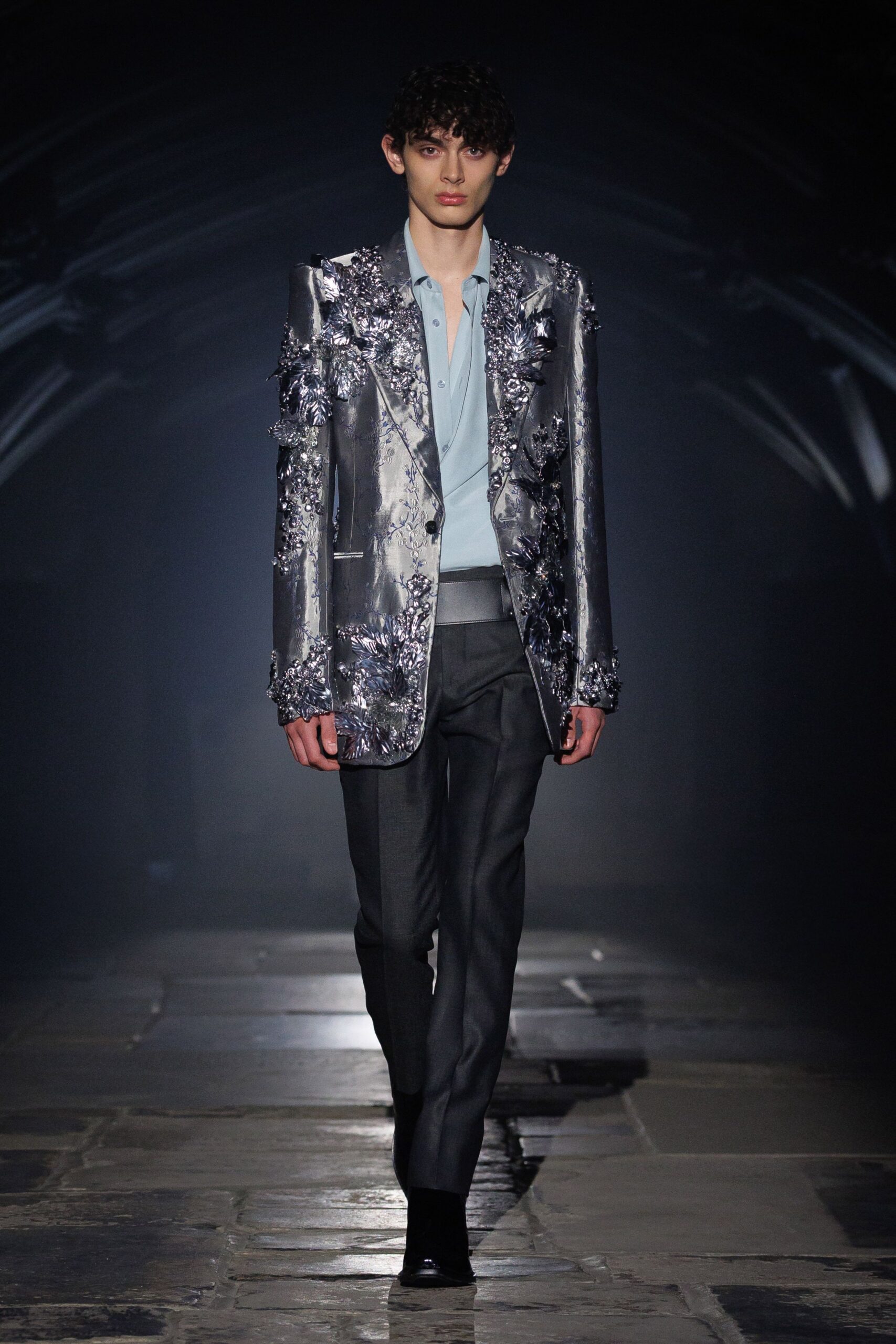 Silver lamé blazer crusted with raised floral appliqué across the shoulders. Powder-blue shirt worn open, a soft fold at the placket. Charcoal tailored trousers with a satin tuxedo waistband. Black boots disappearing under the hem. Crossing a stone courtyard after midnight, boutonniere already crushed.