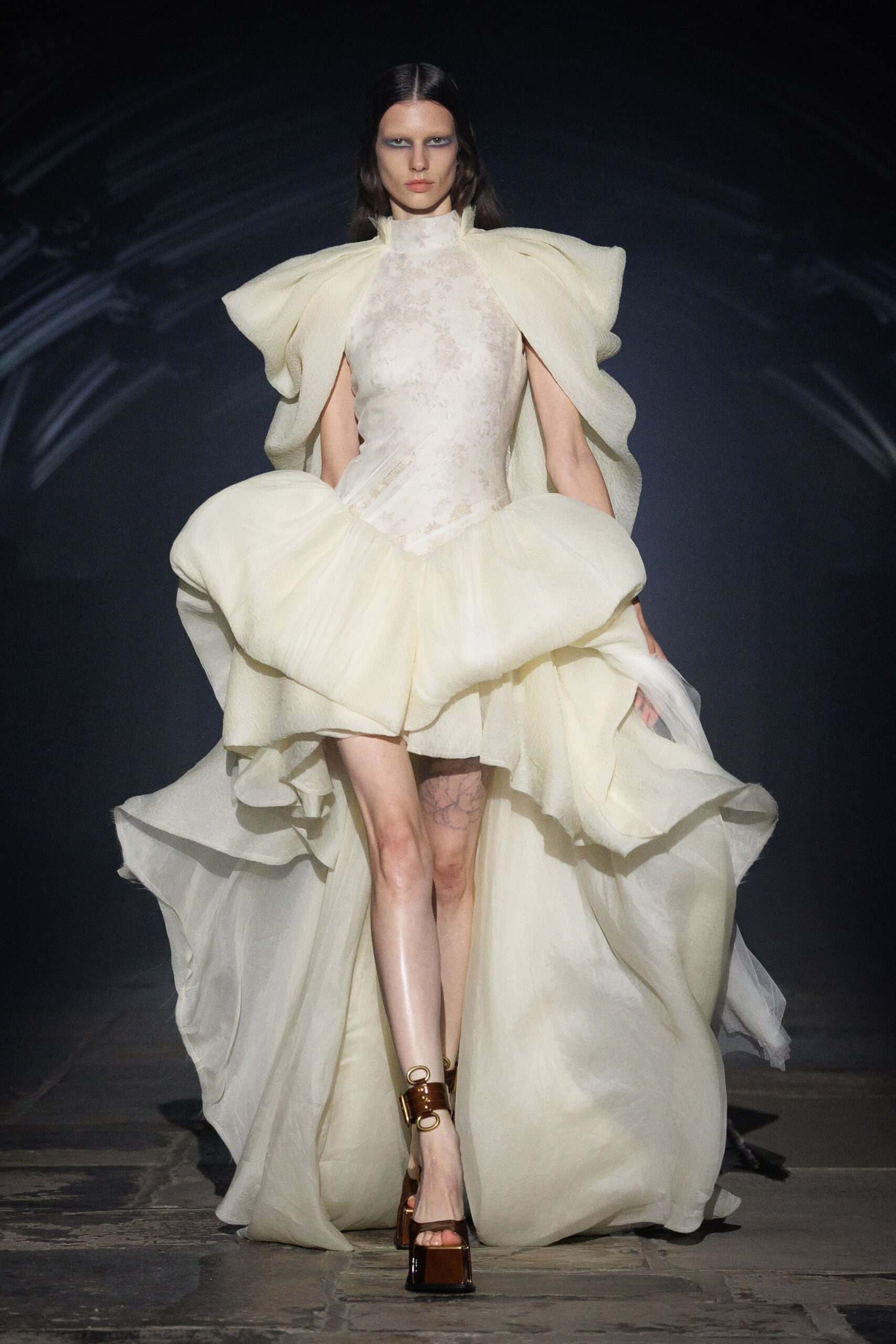 Ivory organza gown with a mottled sheer bodice and inflated ruffle hips. Mahogany patent platforms with oval brass ankle hardware. Crossing a cathedral aisle after the candles have burned low.