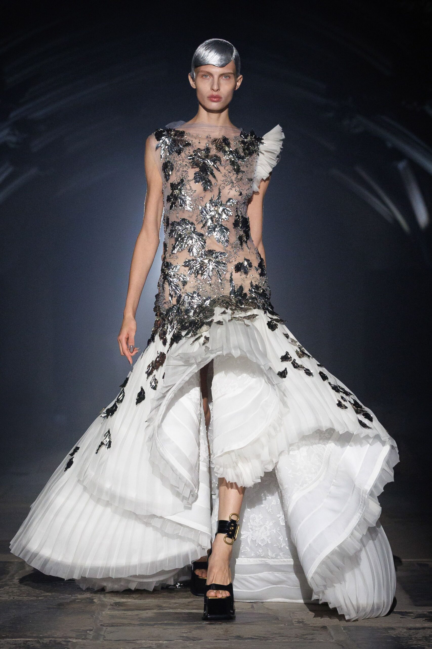 Nude illusion gown crusted with black and silver leaf appliqué. White plissé tiers erupting from the hips into a cathedral sweep. Single white tulle fan at one shoulder. Black patent platforms with a brass ring at the ankle. Crossing a marble hall after the string quartet has packed up.