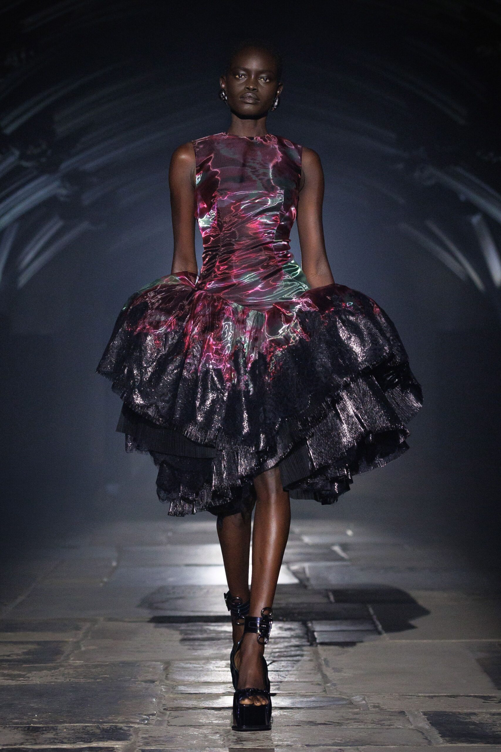 Sleeveless sheath dress in electric magenta marbling, ruched tight through the waist. Black foiled ruffle overskirt, pleated tiers jutting wide from the hips. Black patent platforms with ringed ankle straps. Crossing a stone nave while the organ is still warming up.