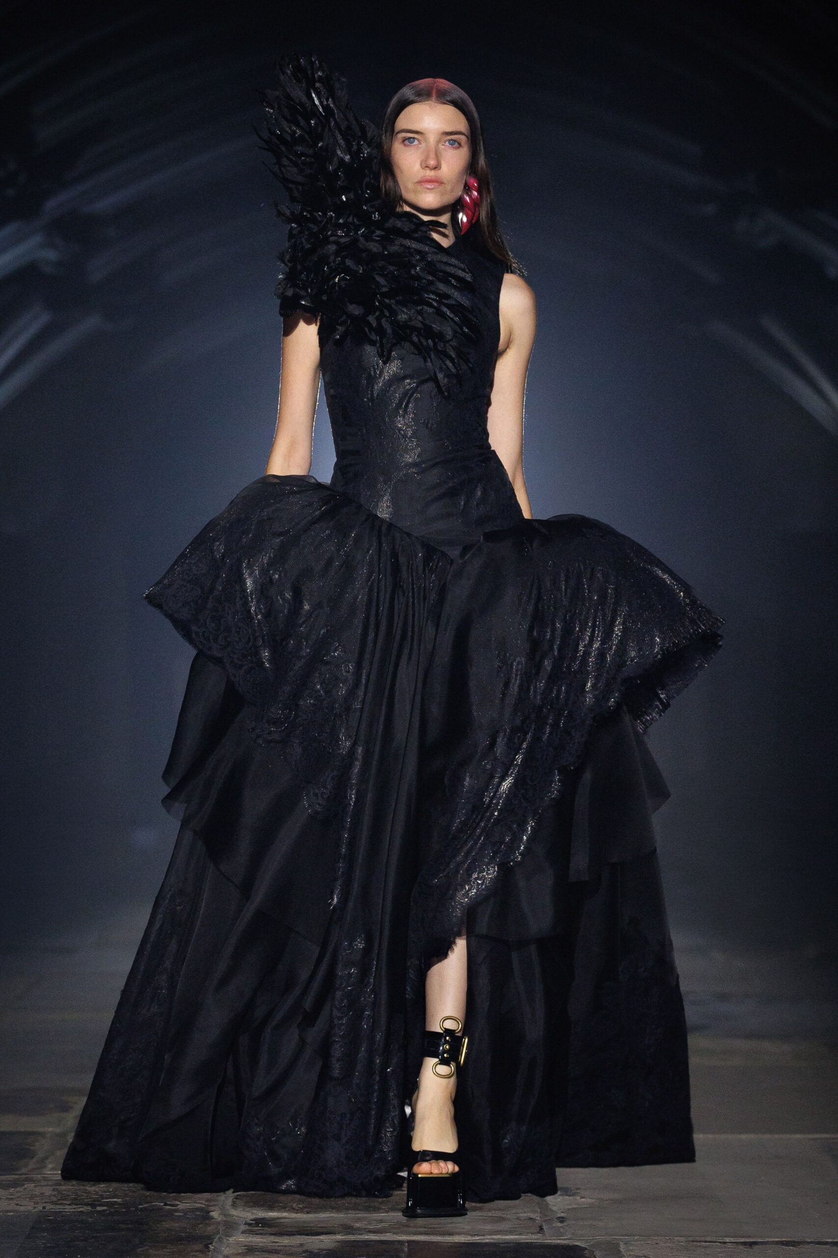 Black evening gown with lacquered feather burst at one shoulder, tiered skirt swelling from the hips. Single cherry-red drop earring. Black platform sandals with oversized gold O-ring ankle cuffs. Crossing a stone cloister after midnight mass let out.