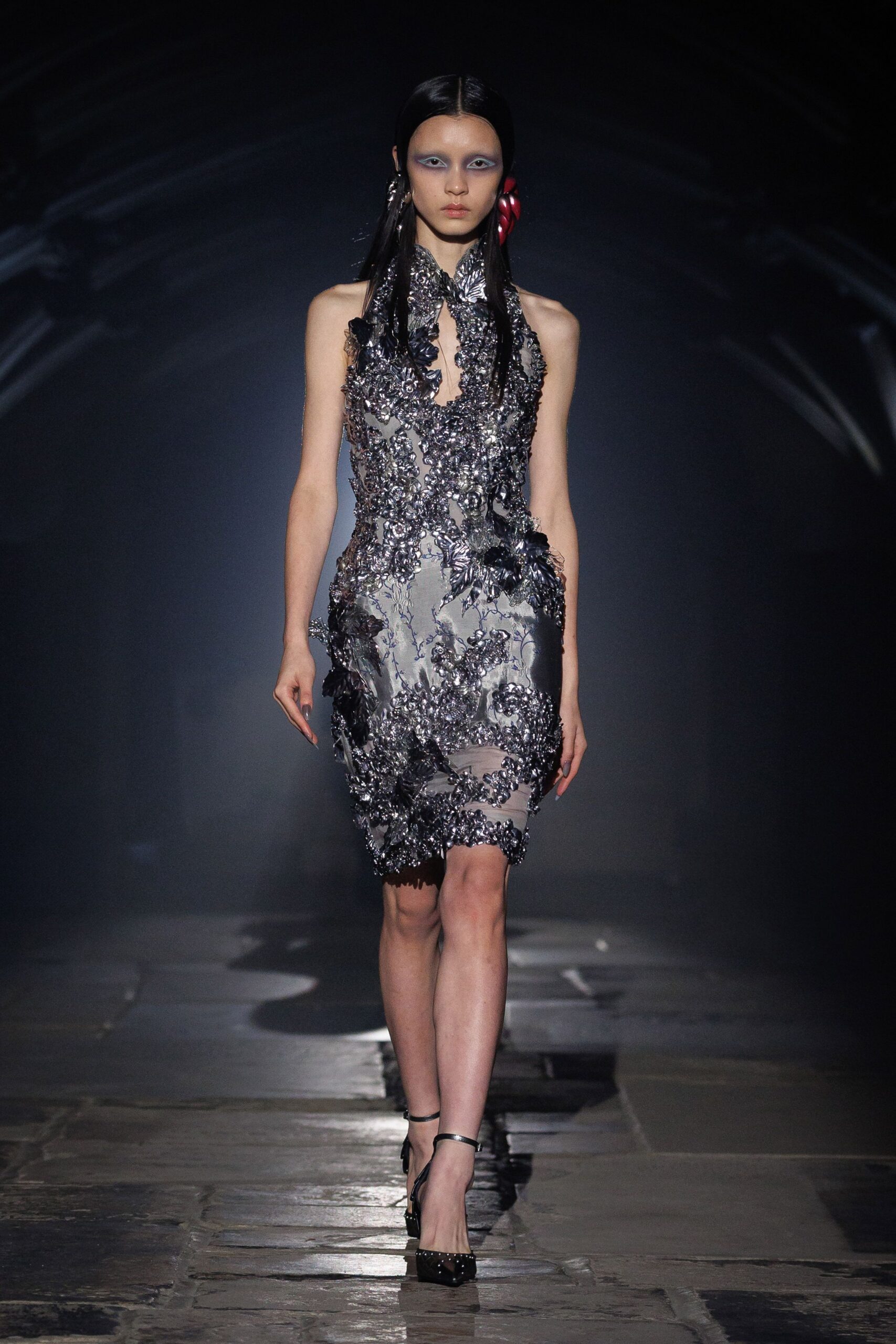 Smoke-silver sheath veiled in raised floral appliqué, cut to a deep keyhole neck. Black ankle-strap pumps with a neat row of studs at the toe. Walking through a conservatory after dark, petals stuck to the damp floor.