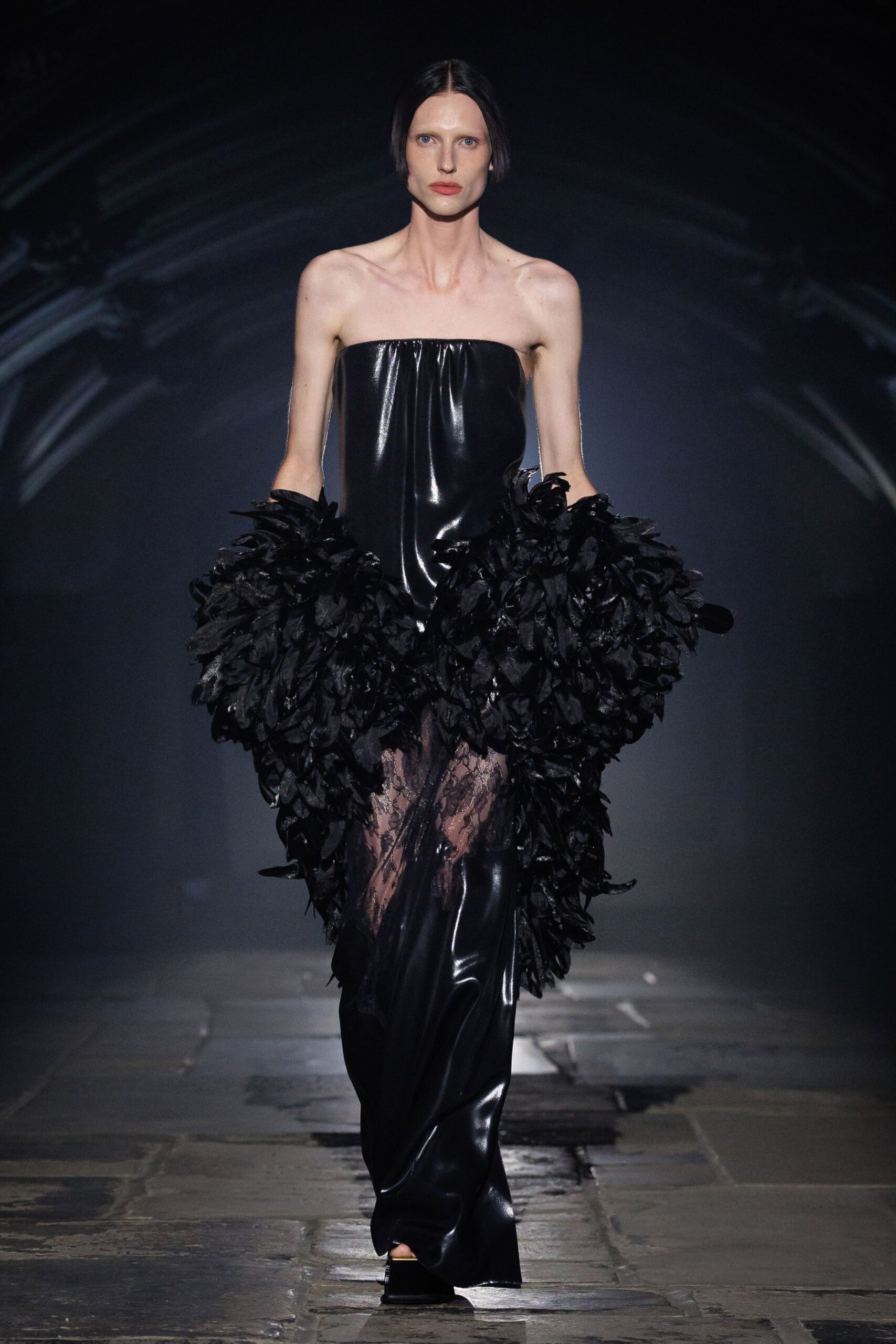 Strapless black vinyl column gown, sides erupting in dense petal-textured flounces. Sheer black lace insert breaking open at the thigh. Square black platform peeking from the dragging hem. Cathedral stones still damp after rain, crossing the nave alone.