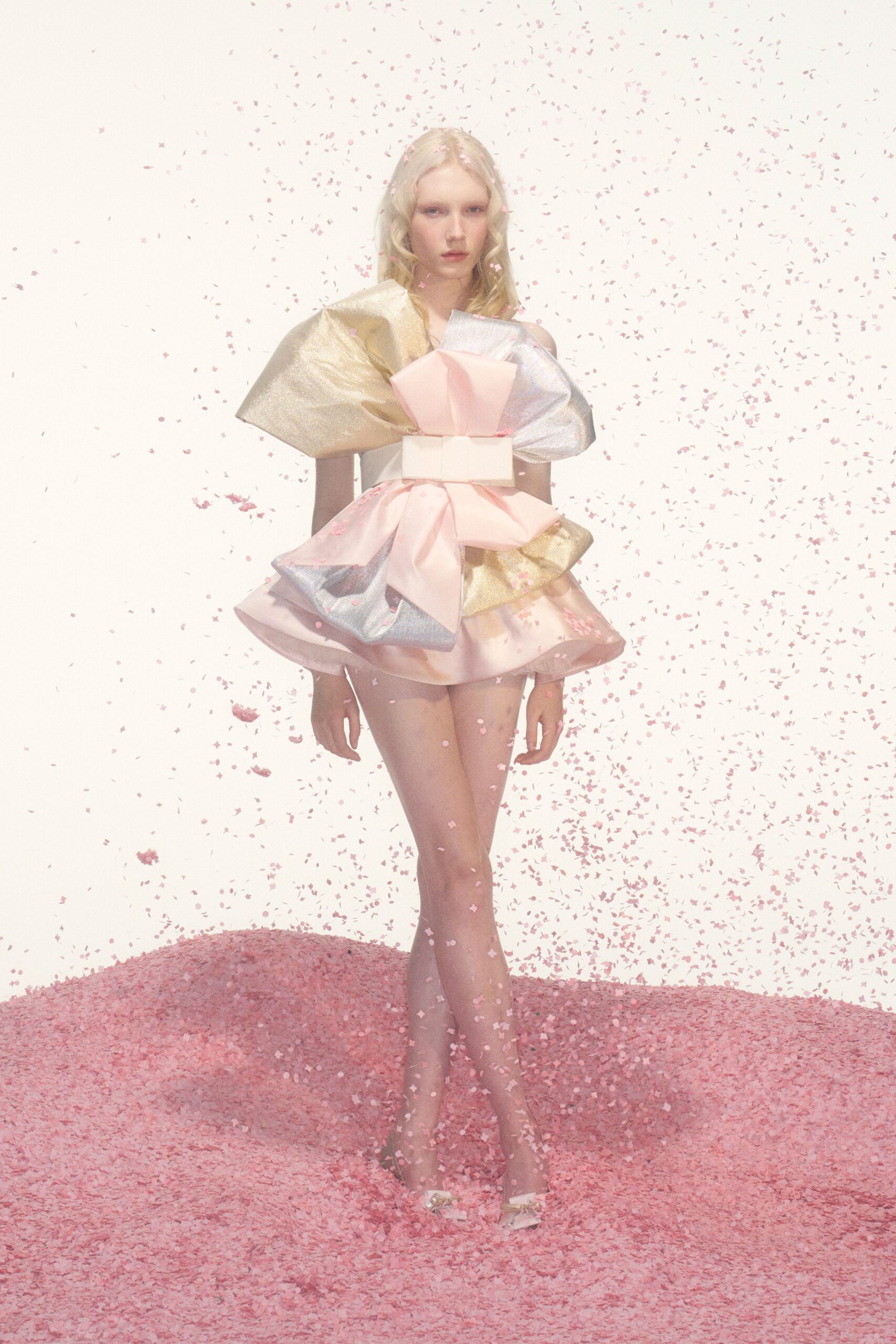 Blush satin mini built from oversized bow folds and metallic lamé panels. Wide ivory waist band cinching the middle like a gift box. Pale pumps topped with crumpled bow ornaments at the toe. Birthday cake table, after the candles, confetti still in your hair.