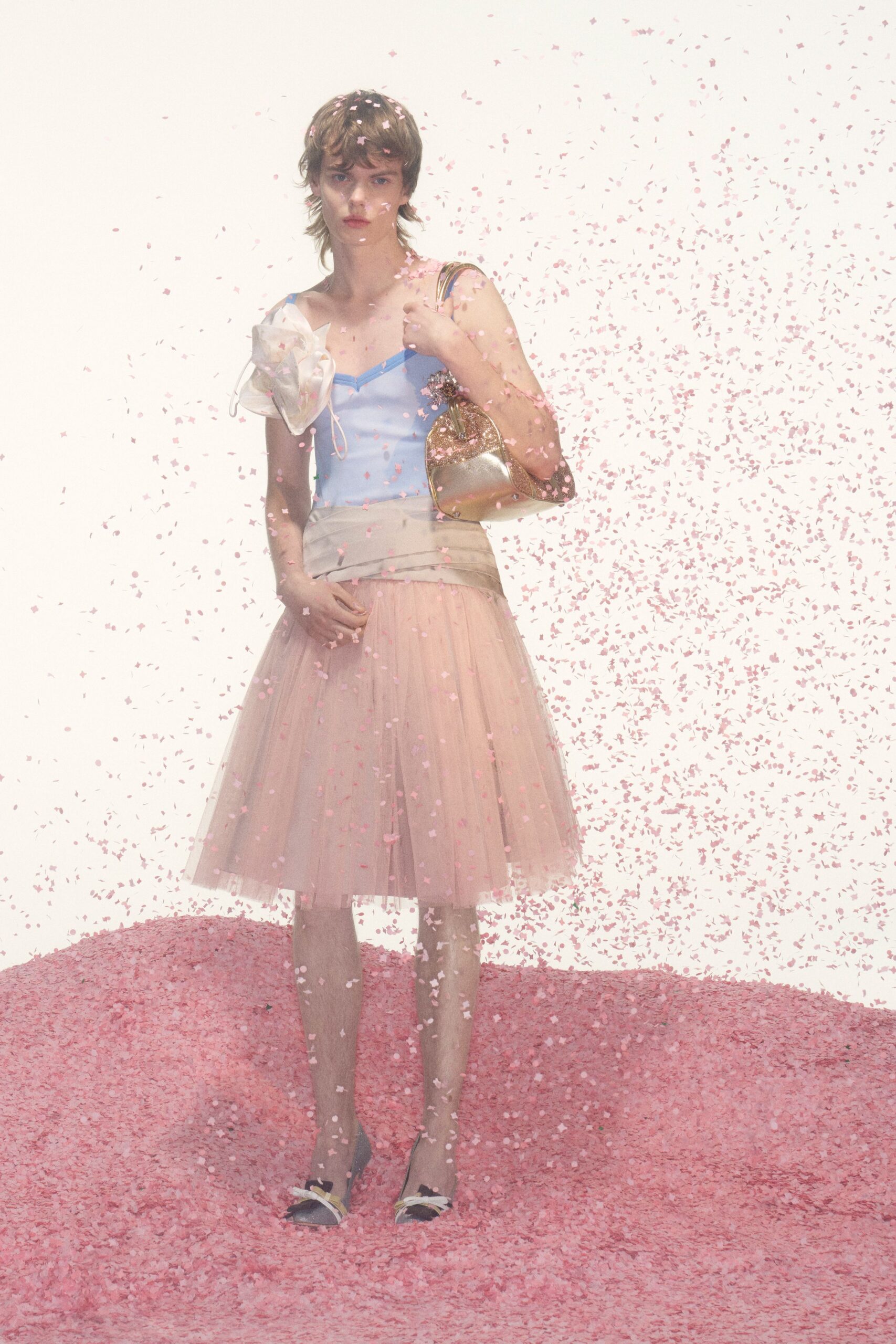 Powder-blue camisole with narrow straps and a sheer organza bloom. Blush tulle skirt under a folded cream hip yoke. Gold shoulder bag, glitter crusting the curved edge. Black flats with pale ribbon bows at the toe. Caught in petal confetti after the town-square wedding lets out.