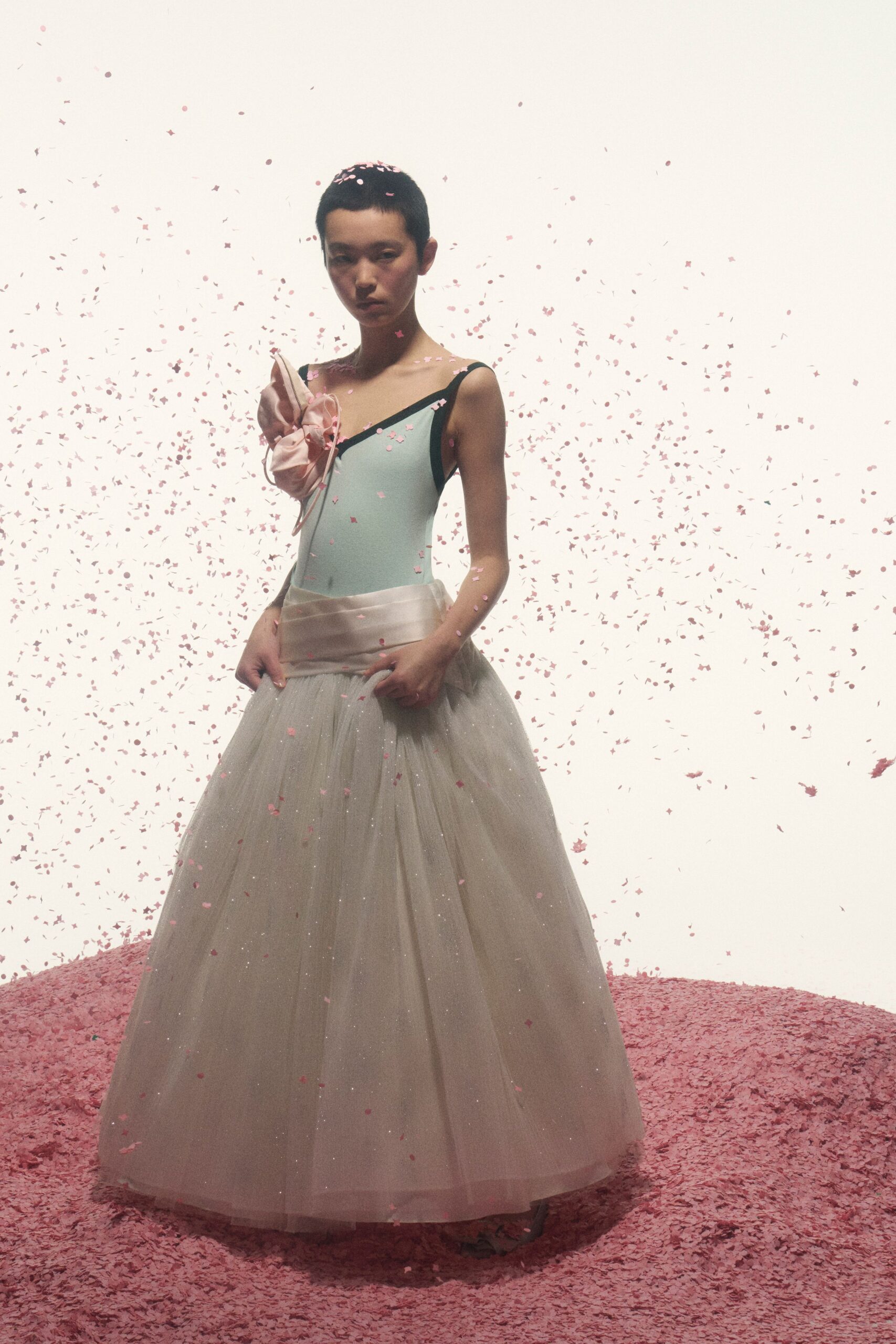 Powder-blue stretch bodice cut on a sharp one-shoulder line. Blush satin rosette pinned high, with a pale ribbon tail. Wide ivory satin sash wrapped low across the hips. Floor-length tulle skirt glittered faintly through its gauzy layers. Confetti still falling after the recital, nobody has started clapping yet.