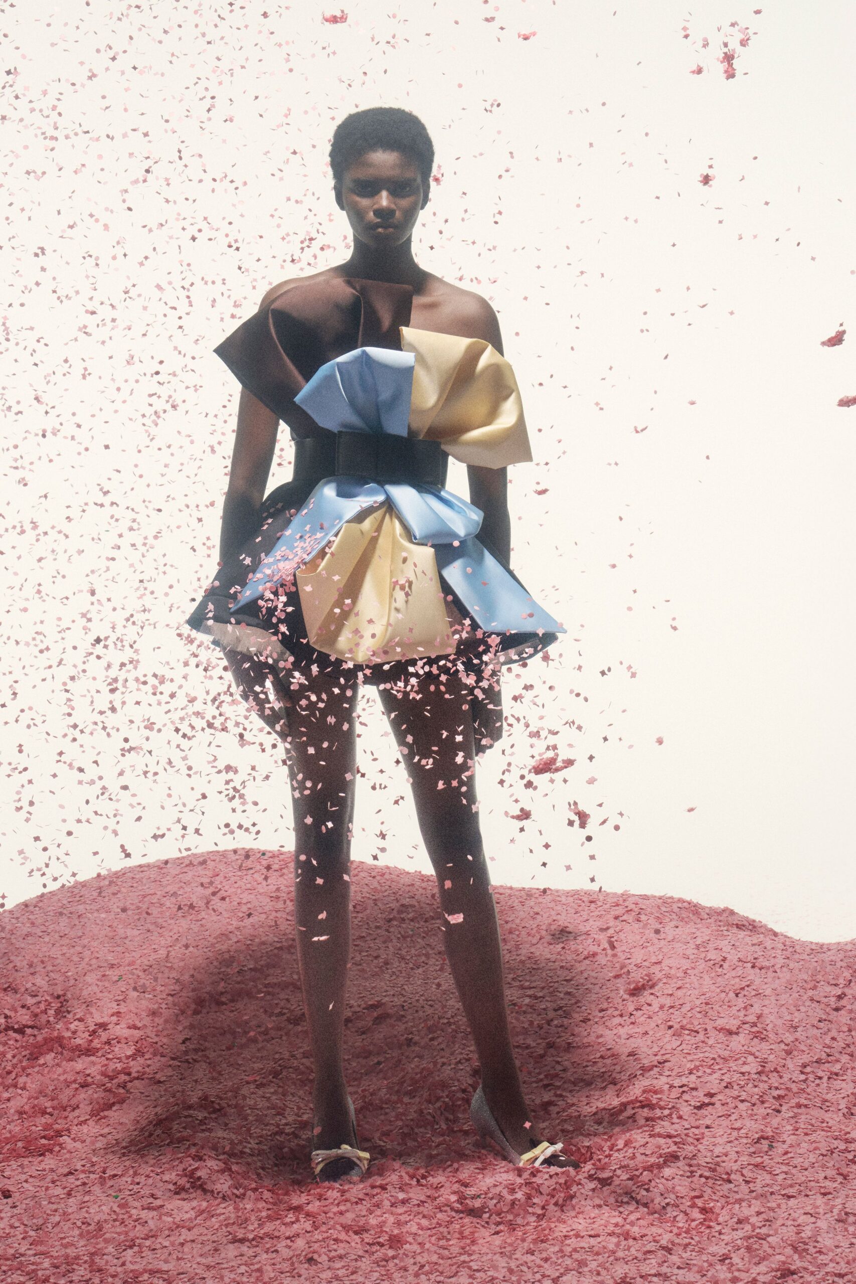 Strapless sculpted mini in chocolate, powder blue and butter satin folds. Wide black waist belt cinching the origami volume hard. Pointed pumps with tiny bow knots at the open toe. Petals in the air, standing on a cake-crumb hillside after the vows.