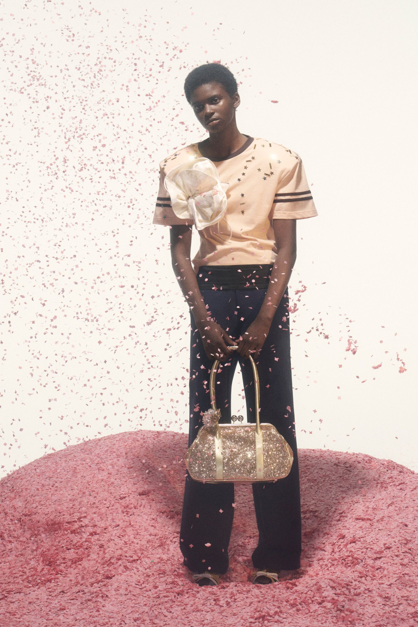 Peach satin tee with black varsity bands and scattered star studs. Oversized ivory organza flower pinned across the chest. Black flared trousers with a wide ribbed waistband. Gold glitter kiss-lock bag with long tubular handles. Pale satin crisscross sandals. Confetti still falling after the school fundraiser ends.