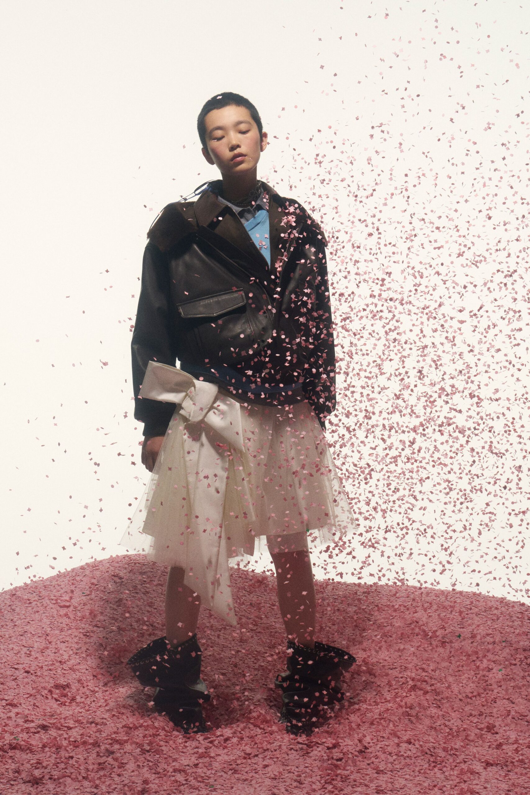 Black leather biker jacket with a dark brown shearling collar panel. Sky-blue V-neck knit layered over a pale shirt collar. Ivory tulle skirt tied with an oversized bow, one tail hanging long. Black slouch boots with silver studs circling the top edge. Cherry blossom confetti sticking to your cuffs after the school recital.