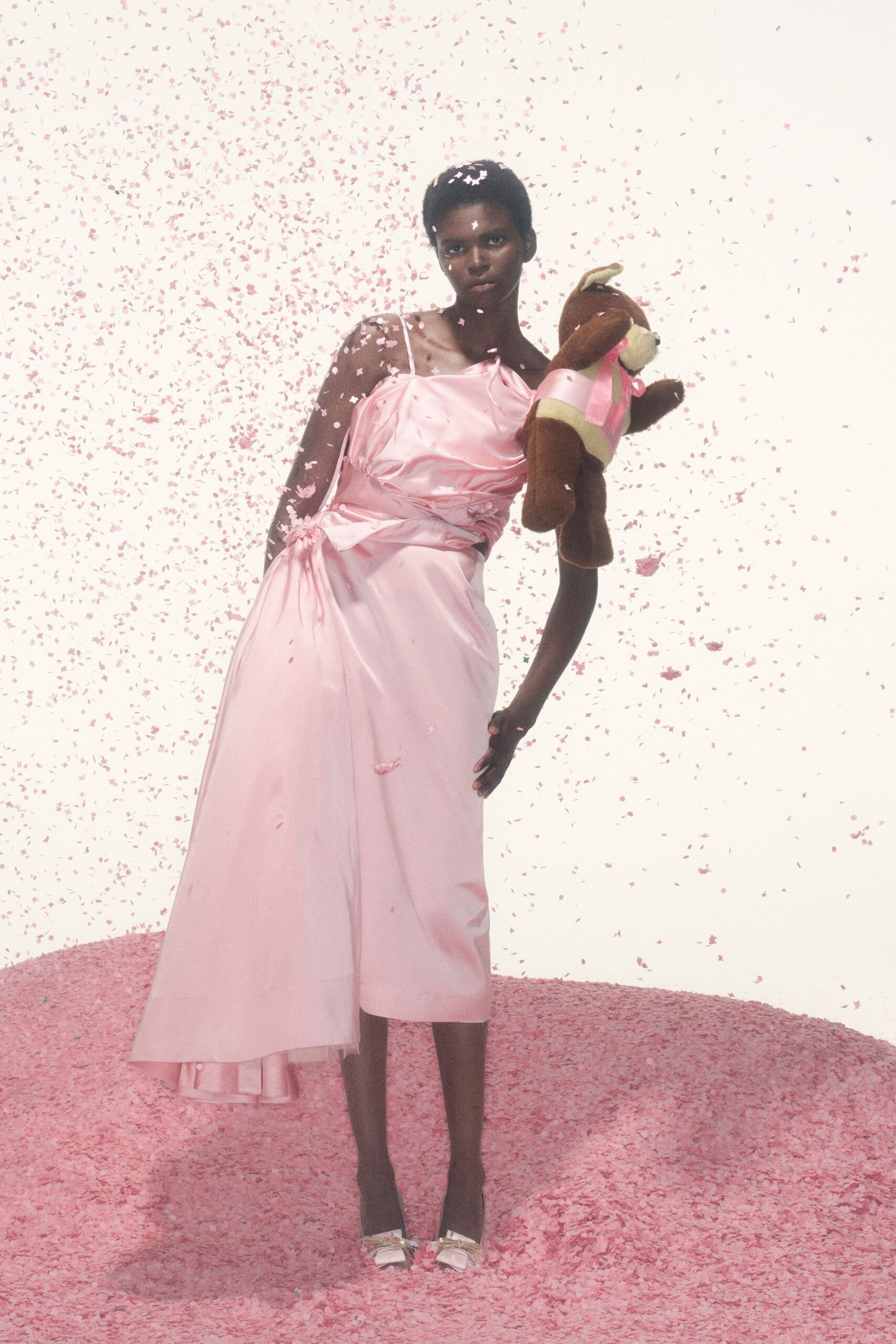 Pink satin slip dress with a draped cowl and tied side sash. Pale satin peep-toes topped with small bow knots. Brown plush bear tucked under the arm in pink ribbons. After the birthday party, carrying home the prize from the game stall.