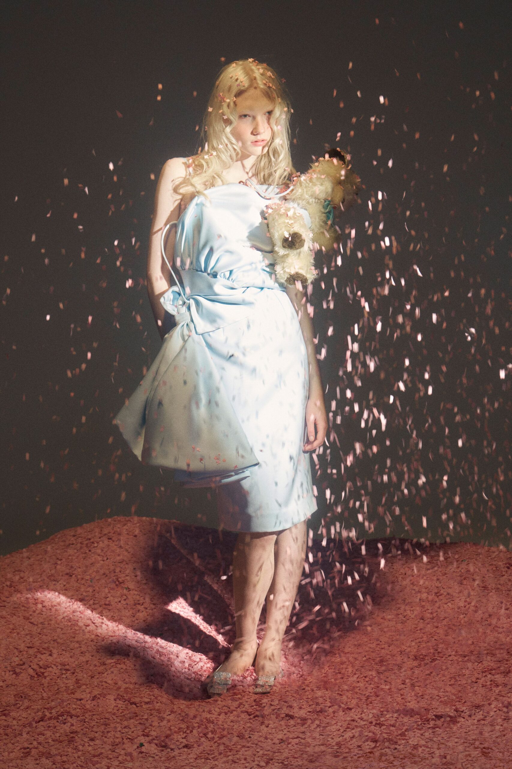 Powder-blue satin slip dress, one strap fallen and a tied pannier jutting sideways. Shaggy cream teddy bear tucked under one arm, turquoise ribbon at the neck. Silver glitter mules with square toes. After the birthday party, standing alone in the confetti drift.
