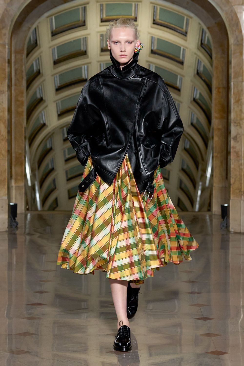 Oversized black leather jacket with a funnel neck and skewed snap placket. Full mid-calf skirt in glossy bias plaid, pooling into deep swinging folds. Black patent lace-ups with a blunt rounded toe. Cluster of bright bead flowers pinned at one ear. Crossing a civic atrium after rain, carrying the whole echo with you.