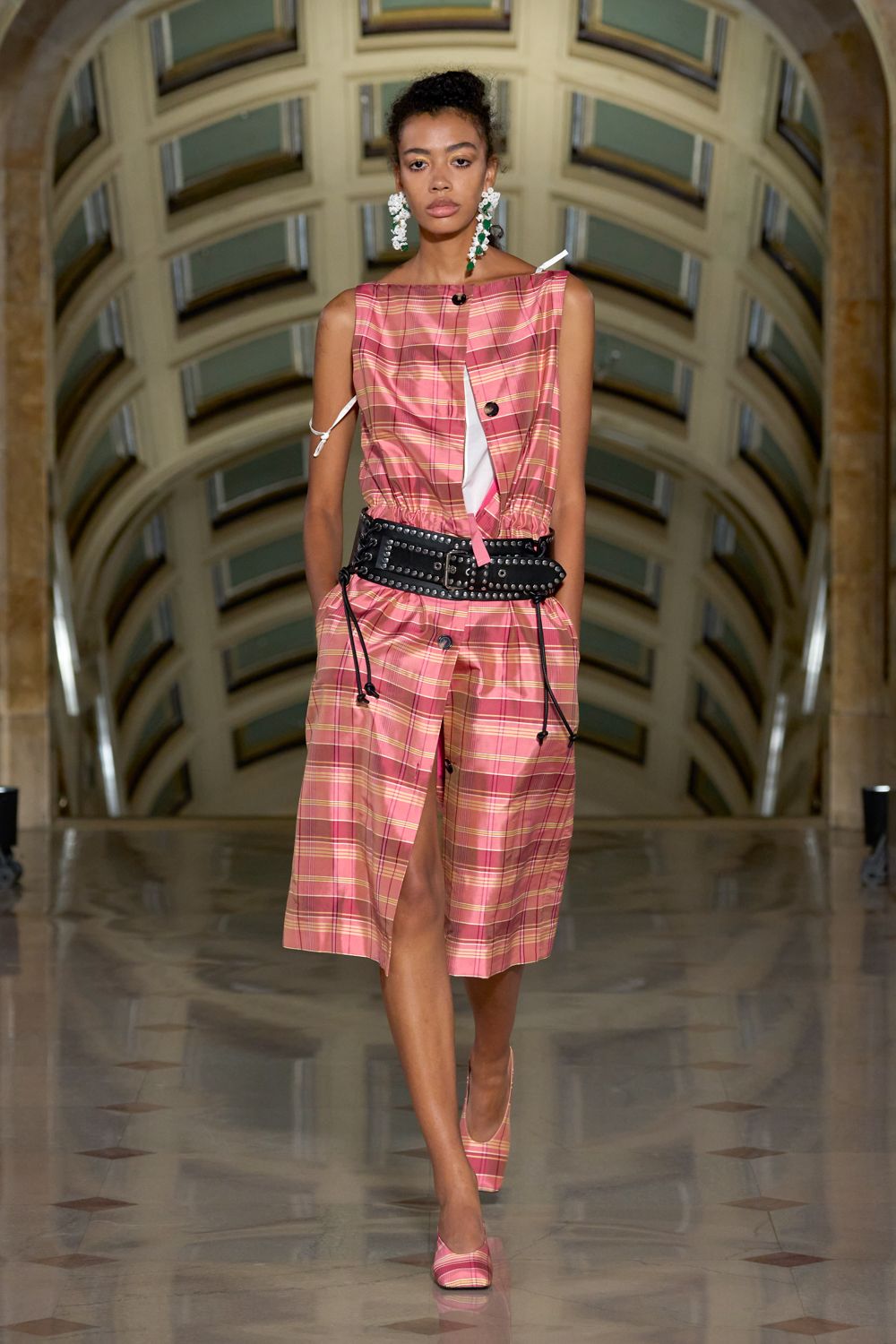 Pink tartan cotton dress with raw armholes, white ties and a split placket. Wide black leather belt packed with silver studs and dangling drawcords. Matching tartan pumps with a sharp closed toe. Long crystal cluster drops with one green stone hanging lower. Crossing a tiled civic hall after fixing your shoes in the taxi.