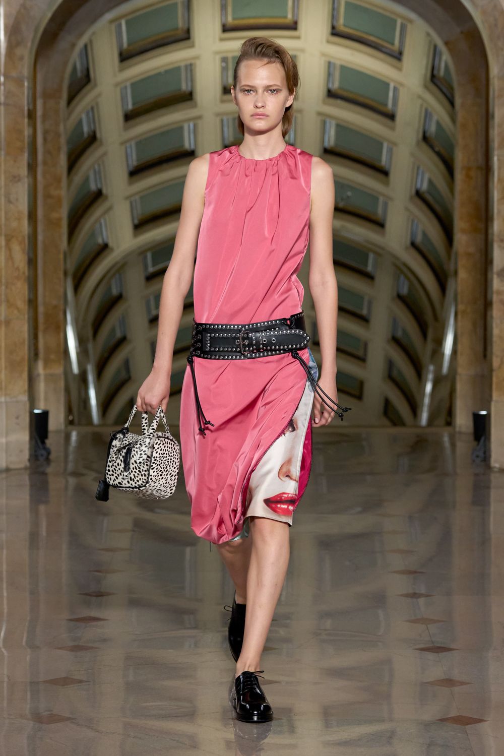 Bubblegum satin dress, gathered neck and a side unzip exposing a face print. Extra-wide black leather belt, ringed with blunt silver studs. Leopard-spotted duffel with chain handles and a swinging tassel. Black patent derbies. Crossing a museum atrium with someone else's overnight bag.