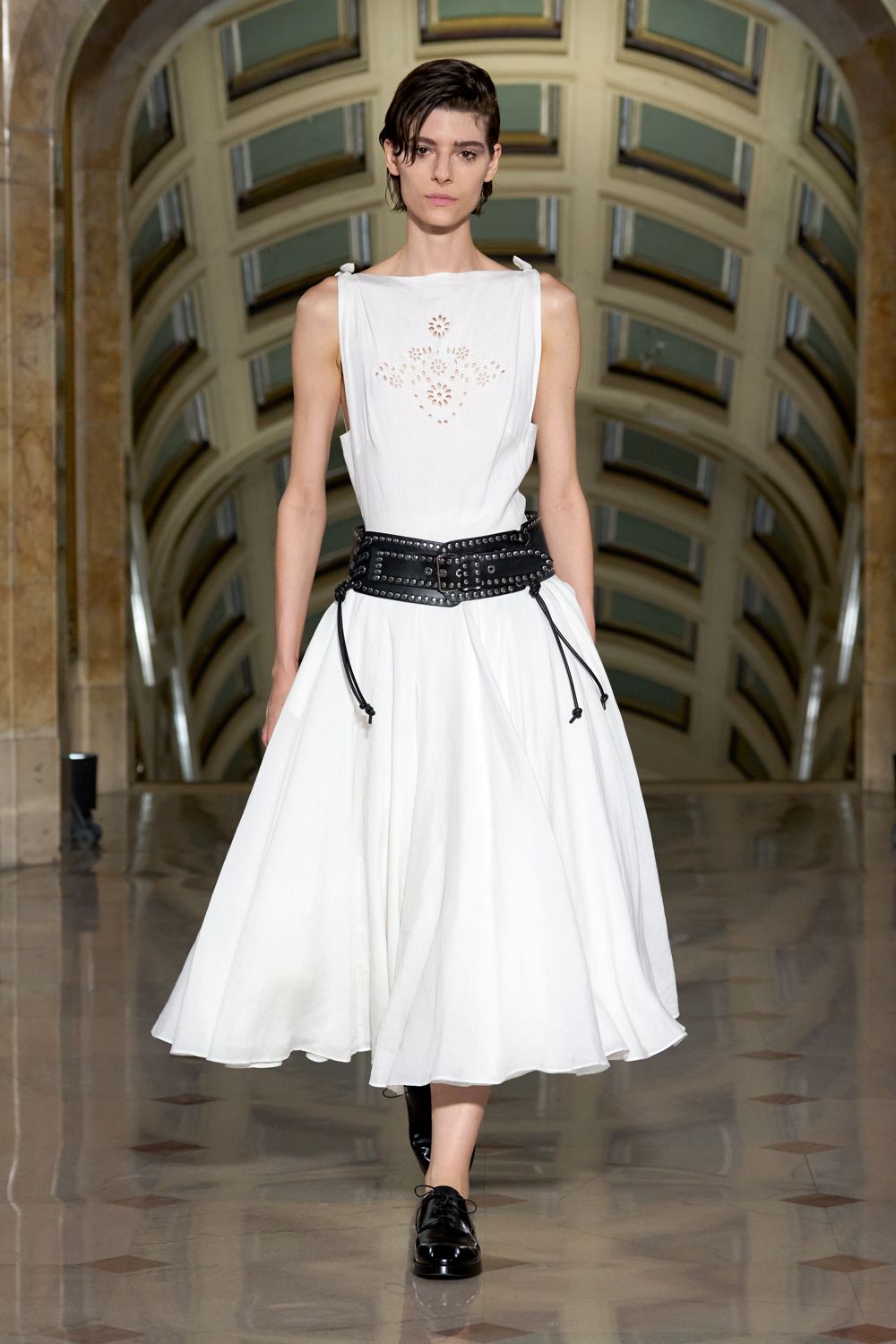 White sleeveless cotton dress, floral eyelets at the chest and a loose gathered skirt. Wide black leather belt studded edge to edge, cords dangling at the hips. Black patent derbies with thin laces. Crossing a museum atrium before the rain starts outside.