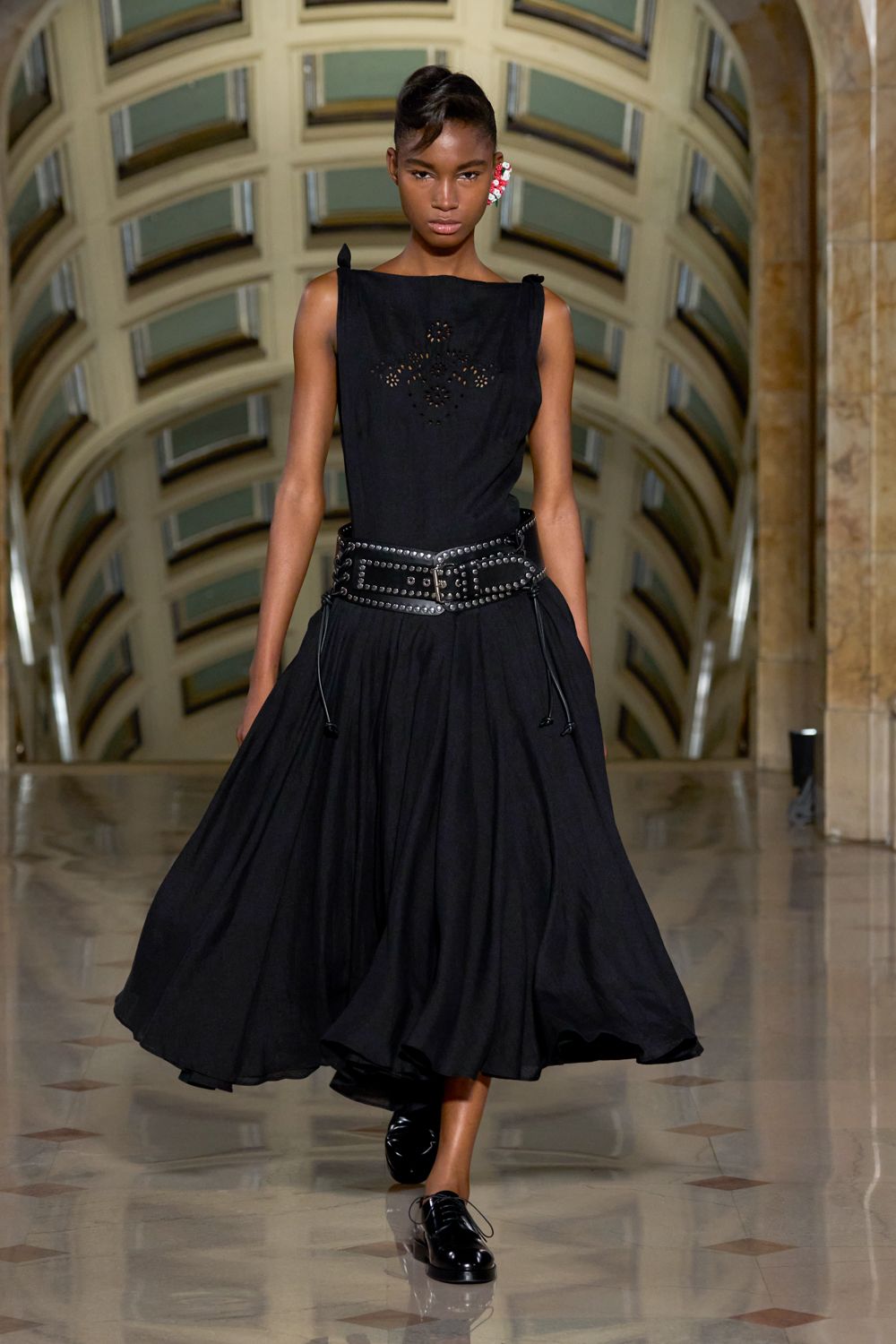 Black sleeveless dress with floral eyelets at the chest and a wide swirling skirt. Black leather belt doubled wide, packed with silver studs and a heavy buckle. Glossy black lace-ups with a blunt rounded toe. Red and white clustered ear cuff. Crossing a museum atrium after rain, marble still giving back the light.