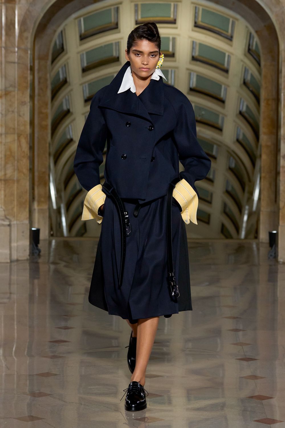Navy double-breasted coat with a broad folded lapel and hanging belt ends. White pointed collar and pale yellow banker cuffs jutting past the sleeves. Dark pleated midi skirt, cut full beneath the cropped coat. Black patent derbies with thin laces. Crossing a marble atrium before the offices open.