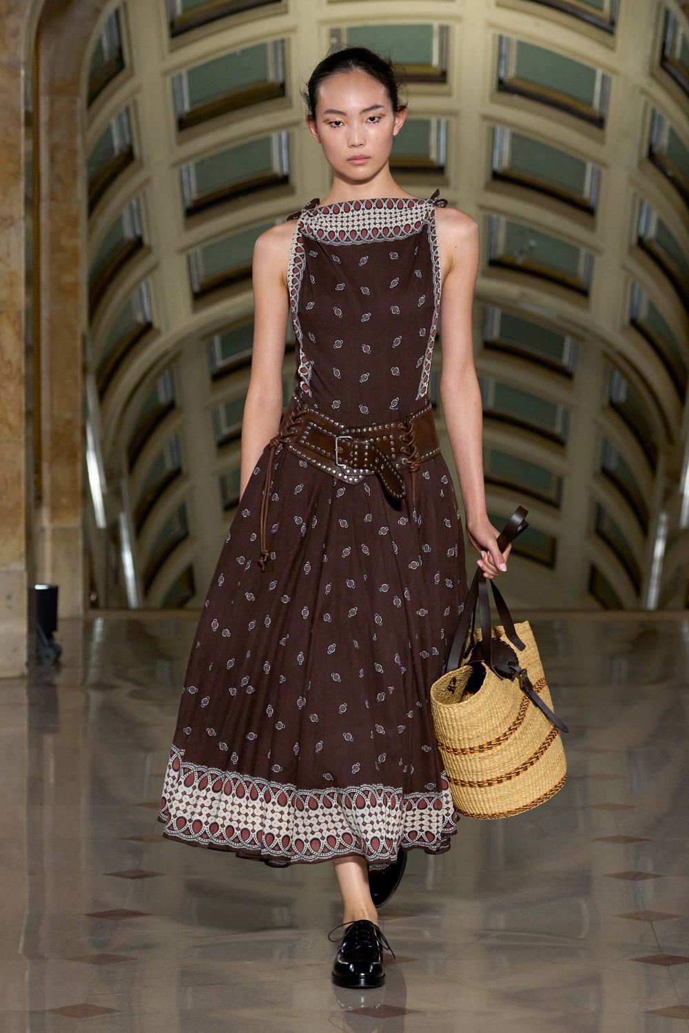 Chocolate sleeveless midi dress with tiny paisley sprigs and a cutwork border. Studded brown leather belt slung low, one lace end dangling. Tall straw basket tote with dark leather straps and braided bands. Black patent derbies with narrow laces. For buying peaches at a covered market before the square heats up.
