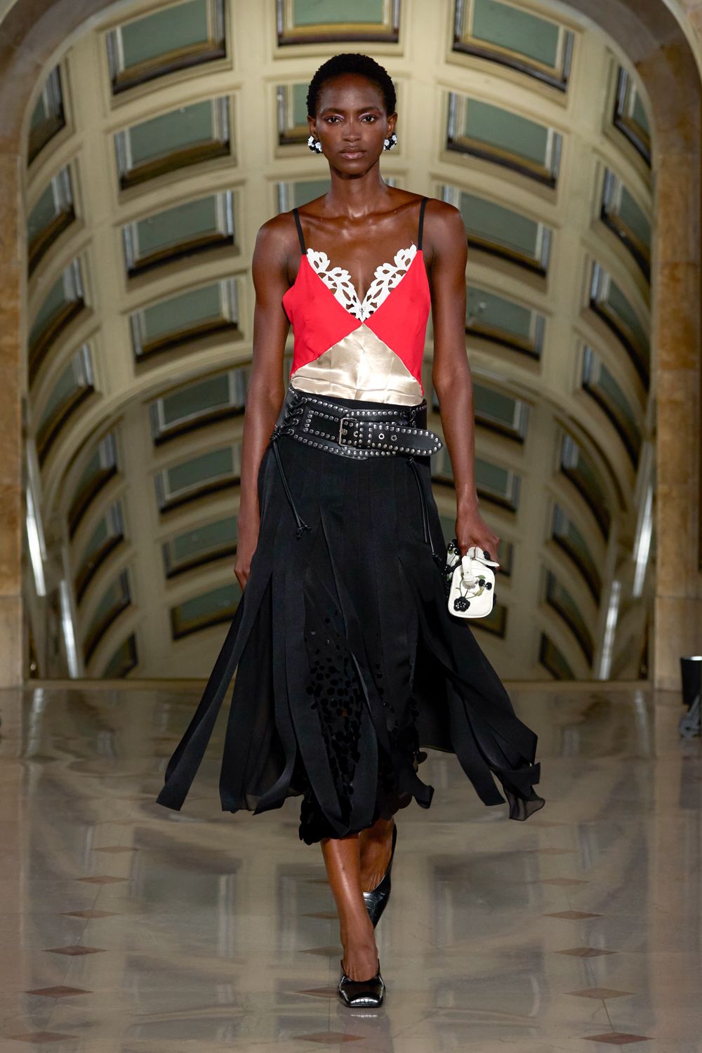Scarlet slip top with black straps, ivory cutwork edging the plunge. Wide black leather belt crowded with silver dome studs. Black godet skirt, laser-cut holes and loose panels whipping outward. Tiny white pouch with black floral appliques. Black patent pumps. Crossing a marble lobby after midnight with the keys in your hand.