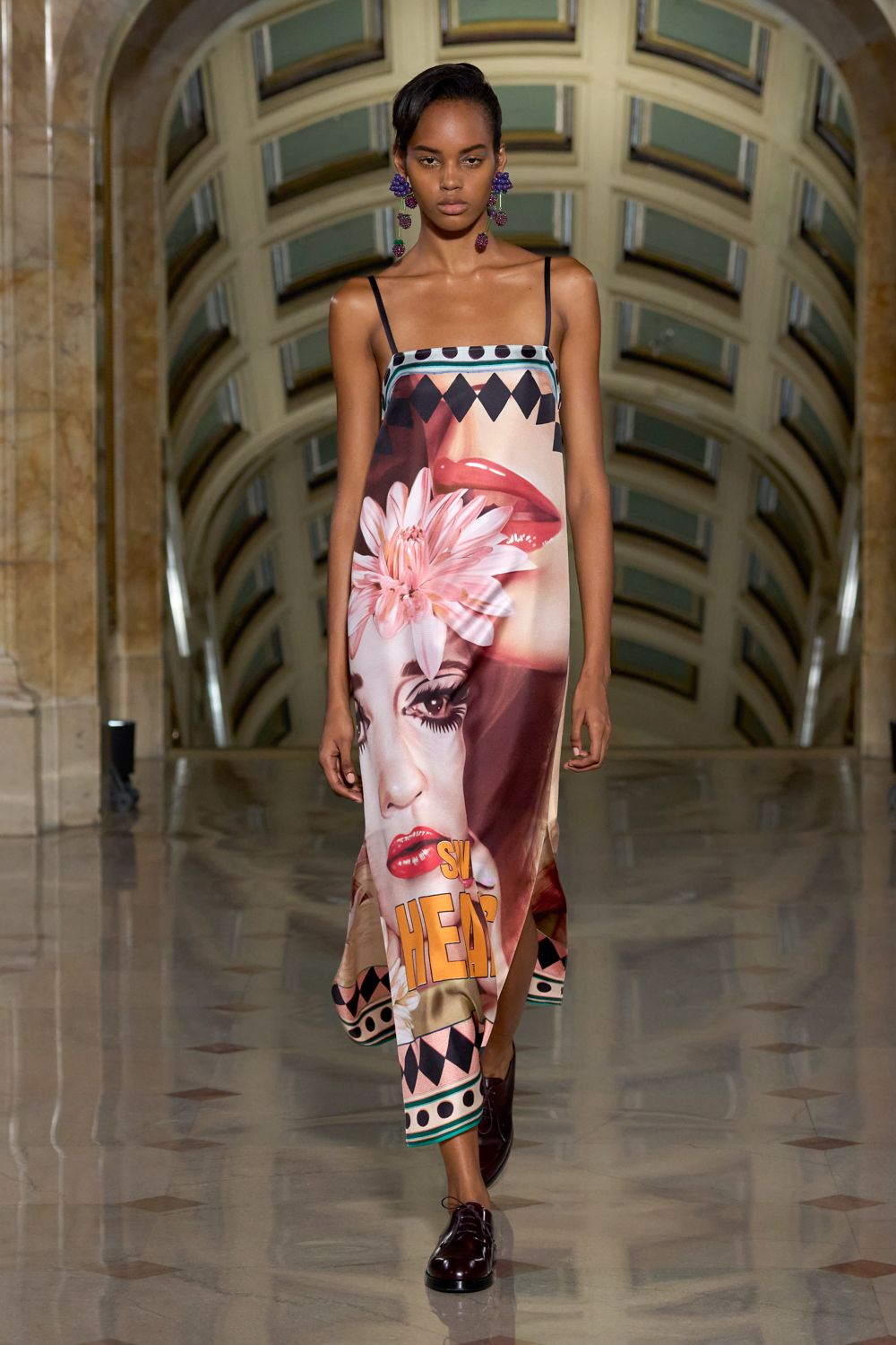 Clustered grape earrings with bead bunches swinging to the shoulders. Column slip in glossy satin, printed with comic-book face and harlequin borders. Oxblood leather derbies with a blunt rounded toe. Crossing a civic hall floor after midnight, still carrying the parade float mood.