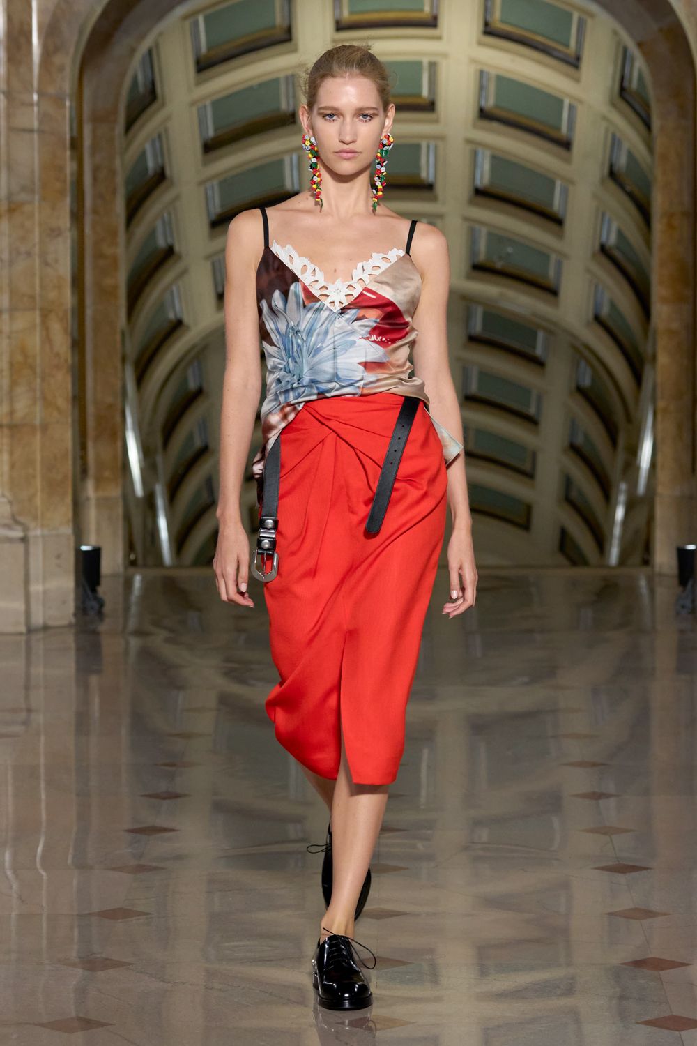 Long clustered bead earrings in candy-bright drops. Silk camisole printed with oversized petals, white cutout lace edging the V. Tomato red wrap skirt, ruched at the hip with one black strap hanging. Black patent derbies with loose laces. Crossing a tiled museum hall after sending the difficult email.